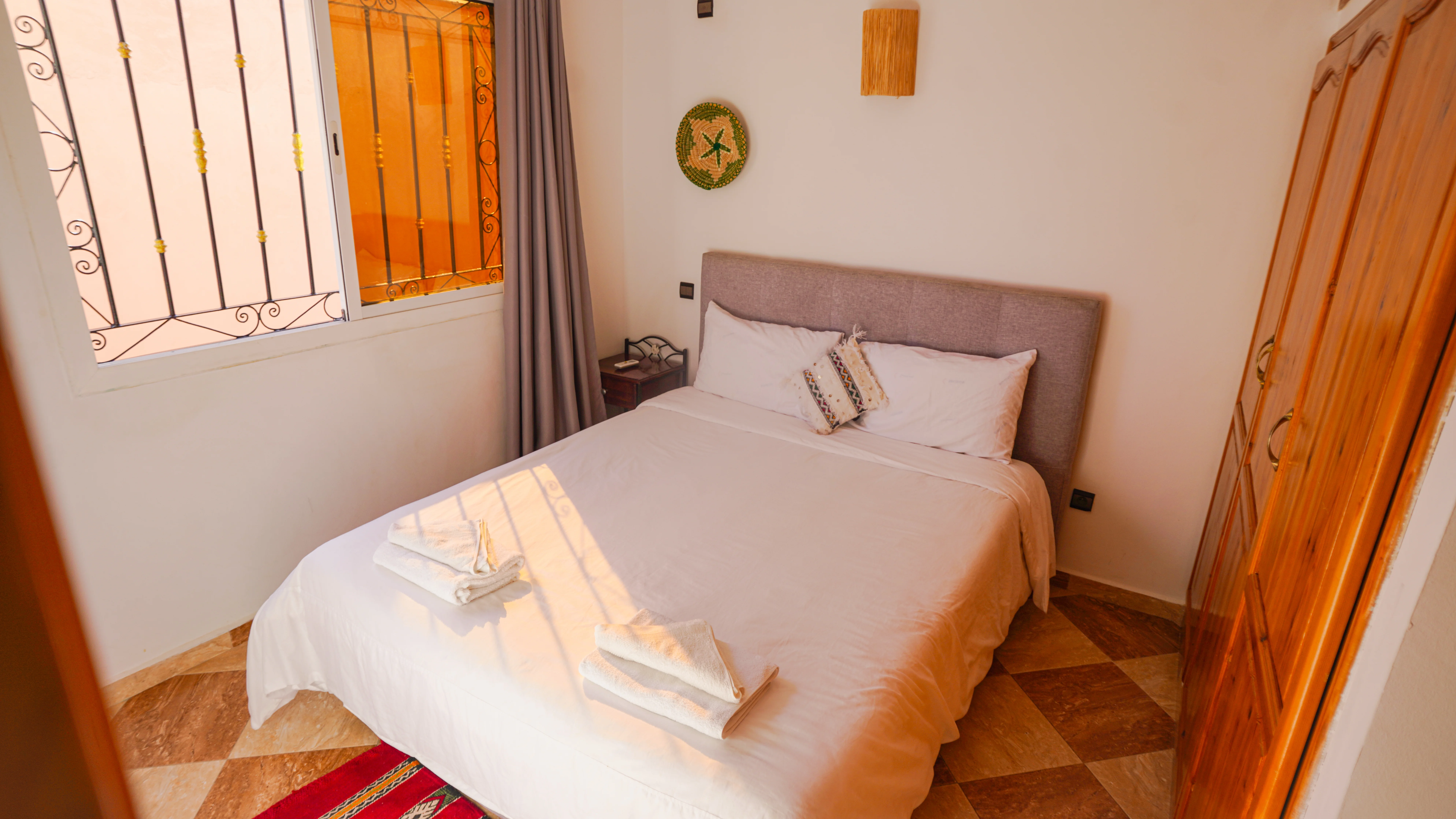 Cozy bedroom with a neatly made bed featuring white linens, two folded towels, decorative pillows, a wooden wardrobe, and a window with wrought iron bars.