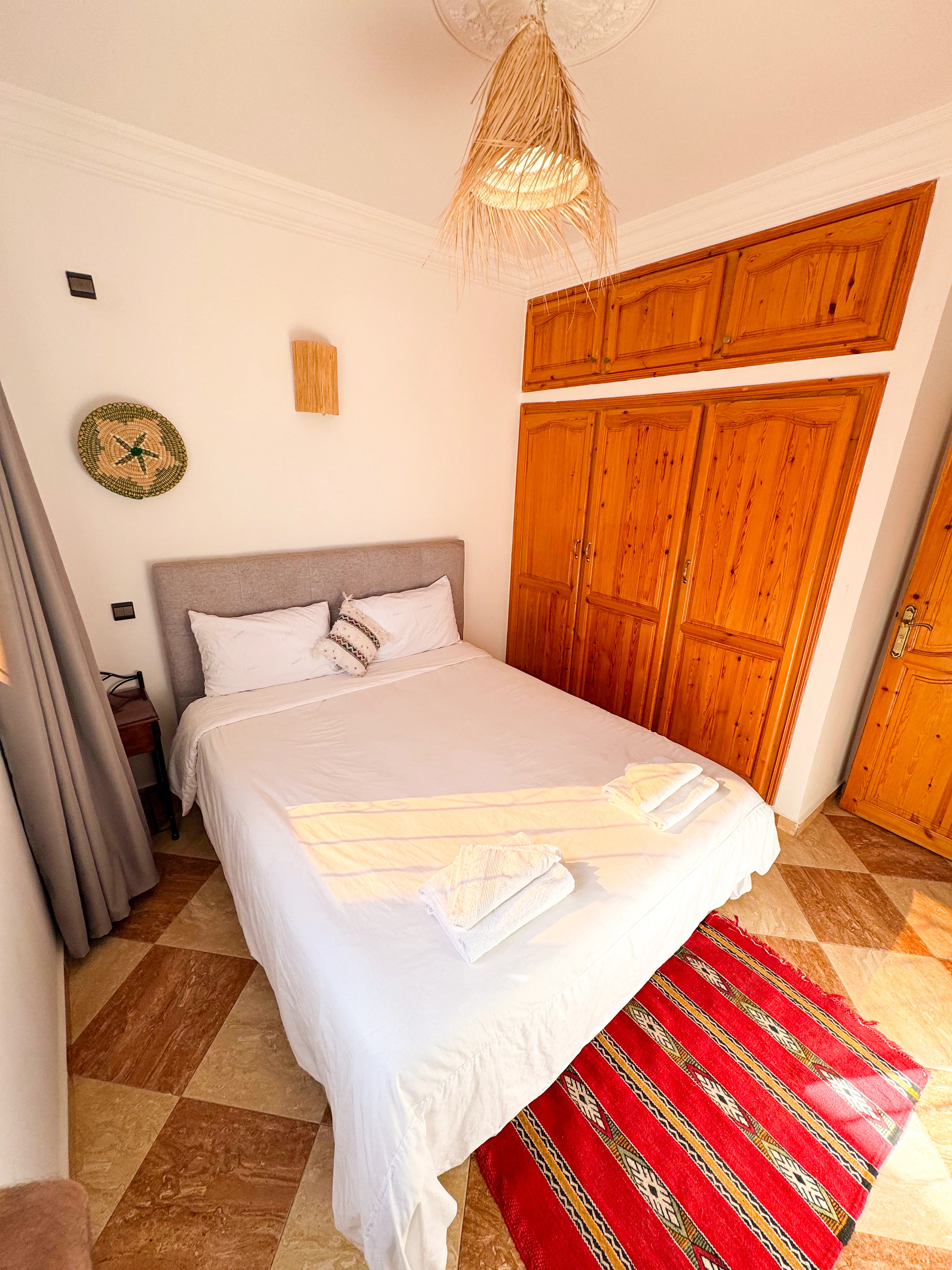 Bright bedroom with a double bed dressed in white linens, wooden wardrobe, red patterned rug, and woven light fixture.