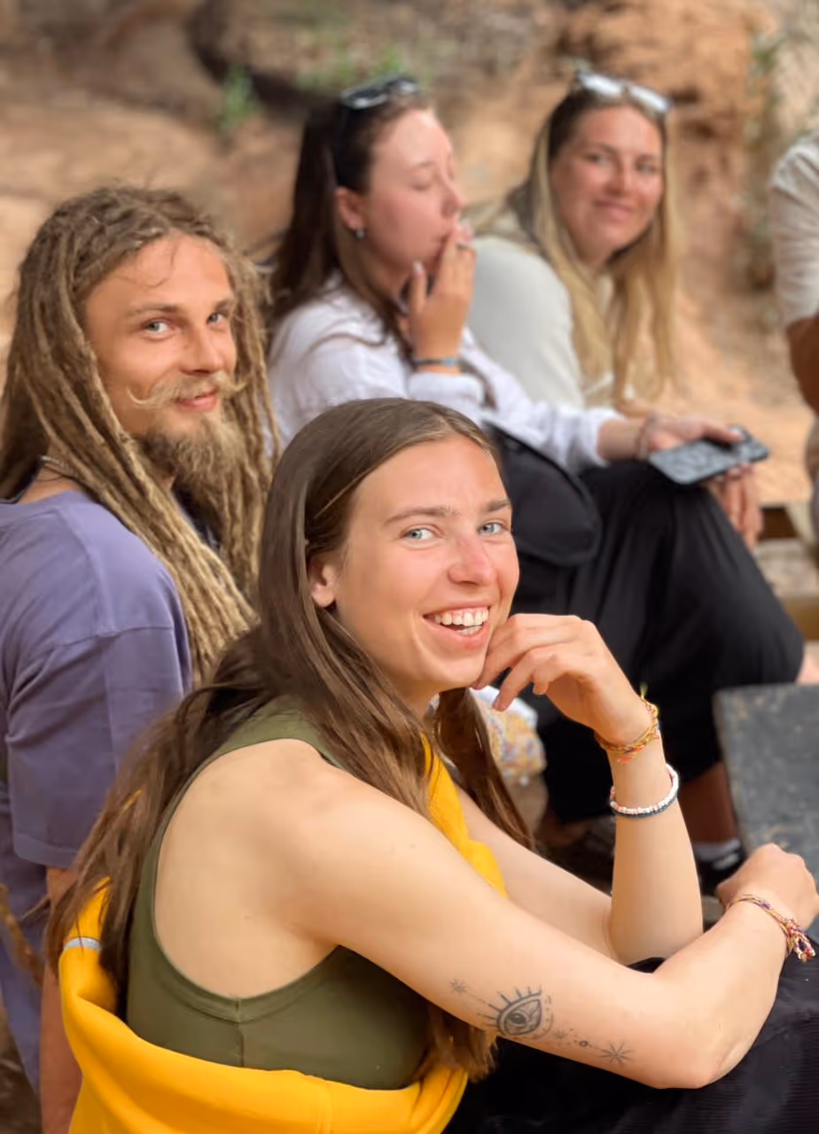 Group of four young adults sitting outdoors, with the front woman smiling and showing a tattoo of an eye on her upper arm.