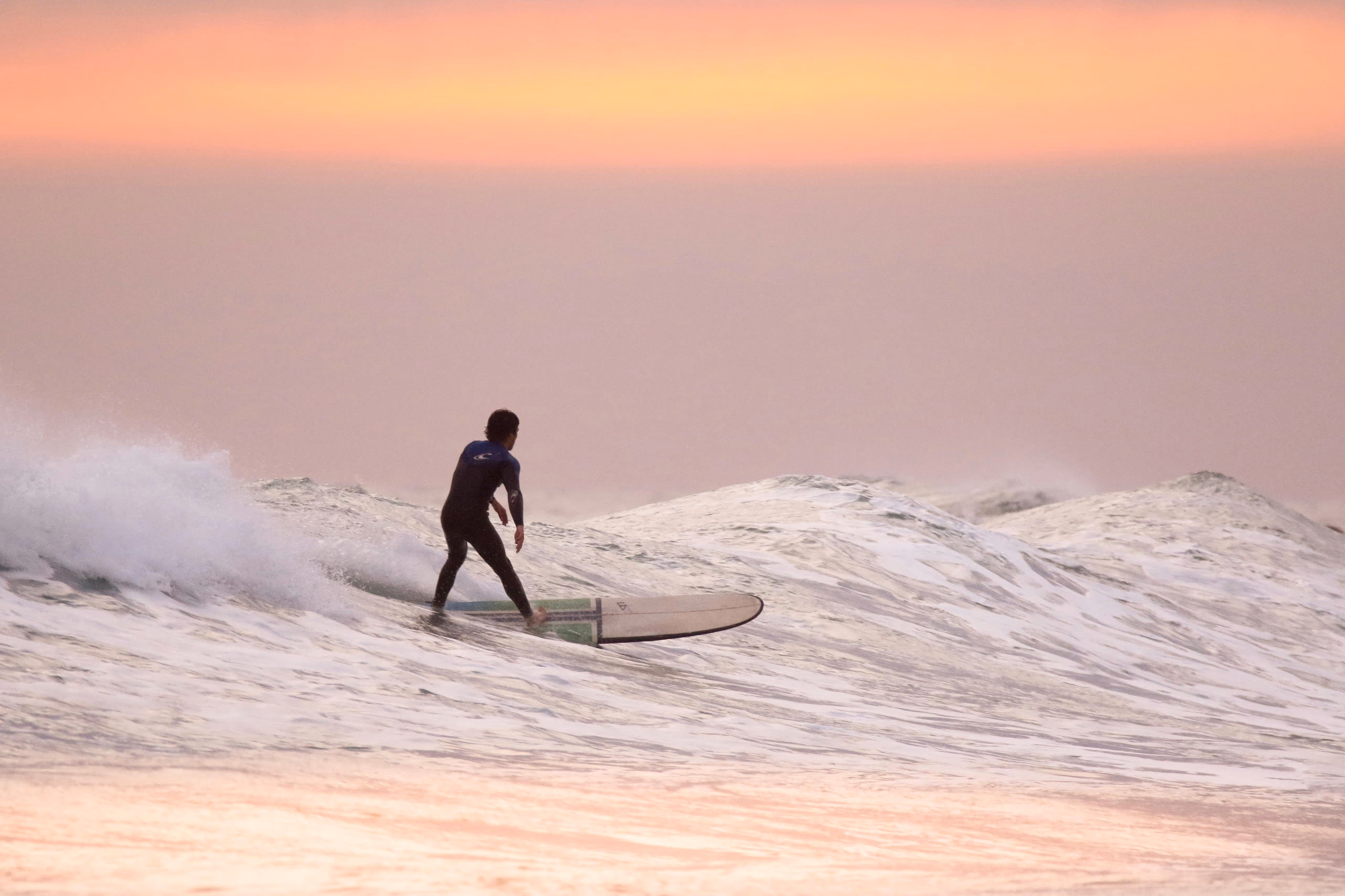 Surfer riding a wave on a longboard at sunset with a pink and orange sky.