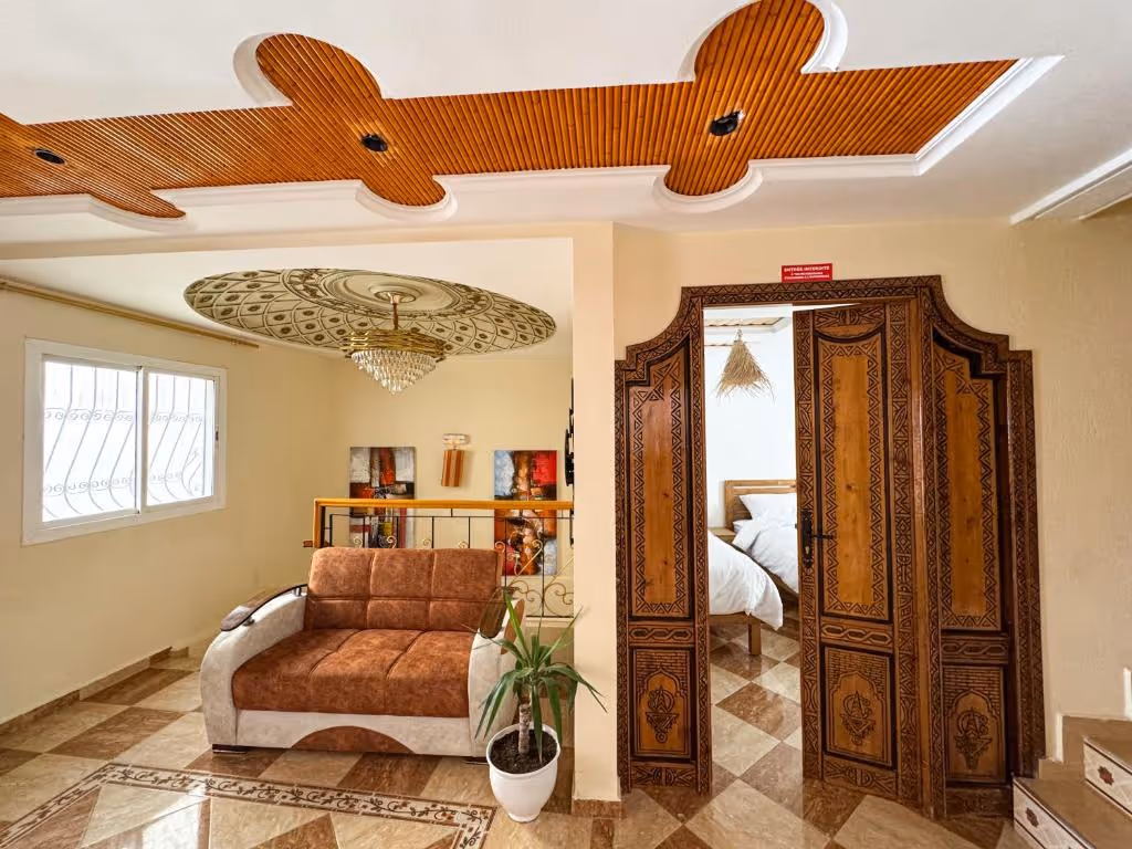 Living space with a brown and beige sofa, decorative wooden ceiling cutouts, a potted plant, and an ornate wooden door leading to a bedroom with white bedding.