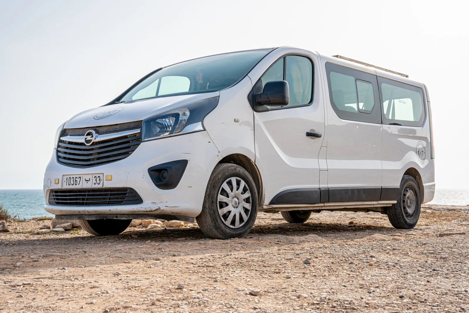 White Opel passenger van parked on rocky ground near a body of water under a clear sky.