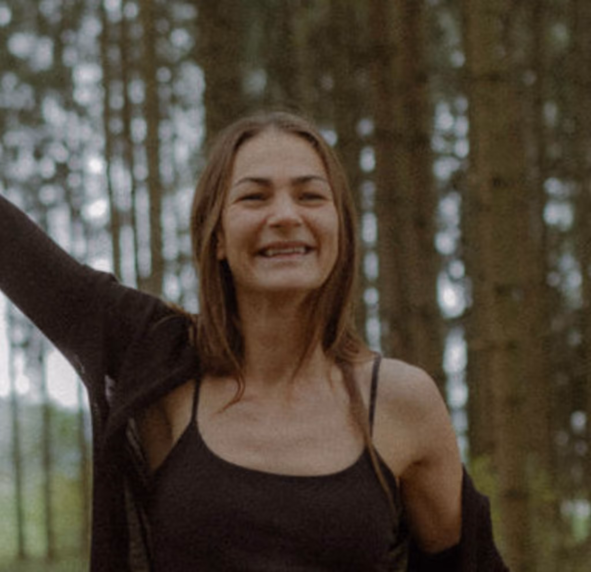 Smiling woman with long brown hair wearing a black tank top and jacket in a forest setting.
