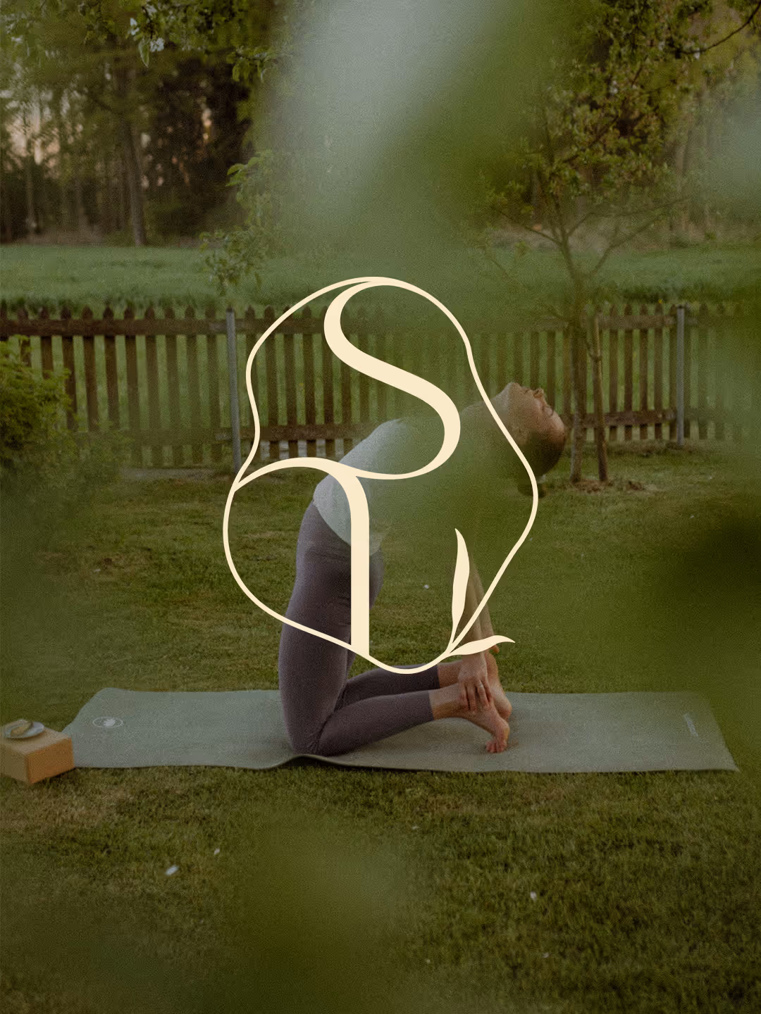 Person performing a yoga backbend pose on a mat outdoors in a grassy area with a wooden fence and trees in the background.
