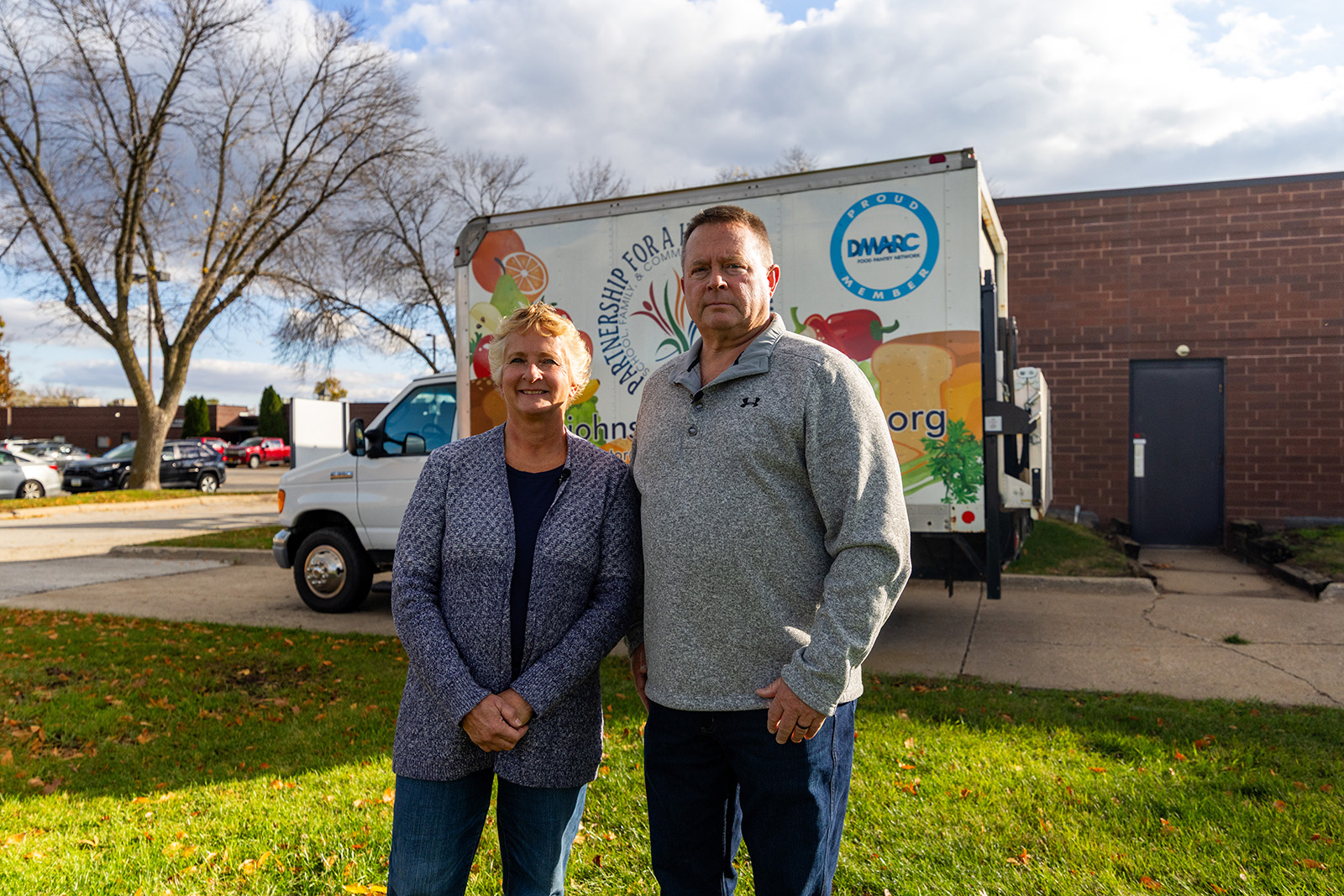 A man and a woman standing on grass in front of a delivery truck with a bright sky and bare trees in the background.