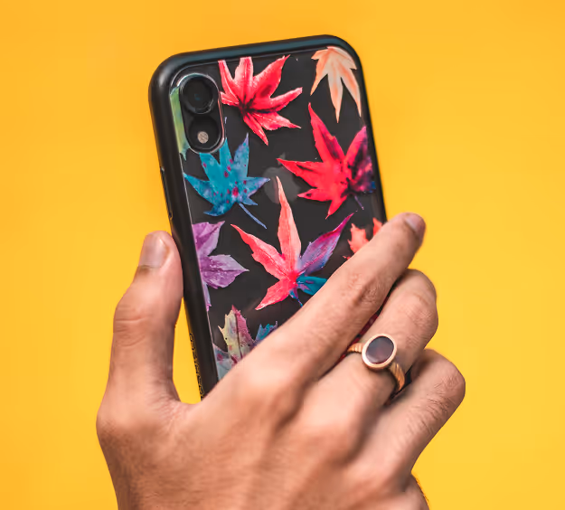 Hand holding a smartphone with a black case decorated with colorful autumn leaves against a yellow background.
