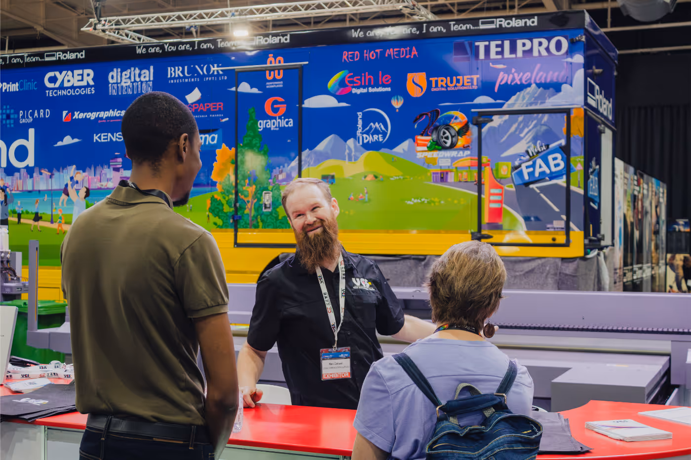 A bearded exhibitor with a badge talks smilingly to two visitors at a colorful trade show booth with various company logos on a blue and green backdrop.