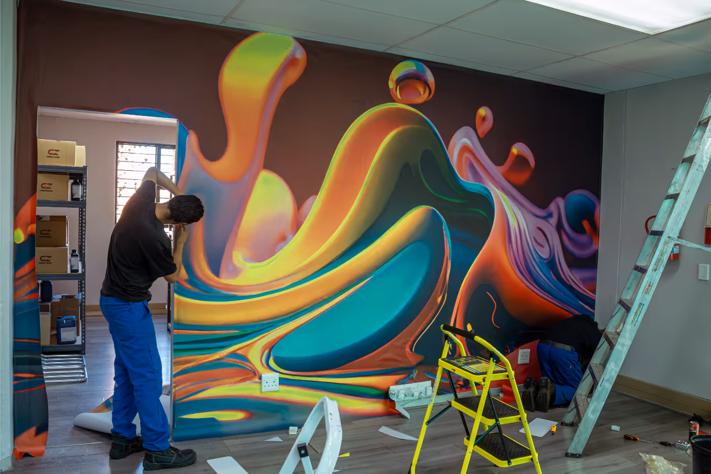 Two workers installing a large colorful abstract mural on an office wall, with ladders and tools nearby.