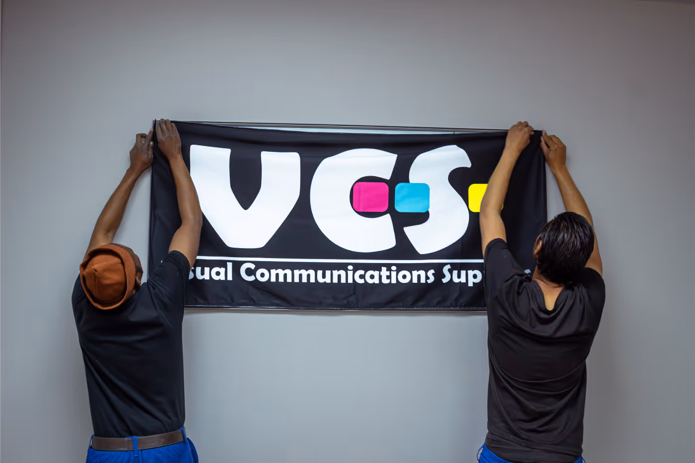 Two men hanging a black banner with large white letters 'VCS' and colorful blocks on a gray wall.