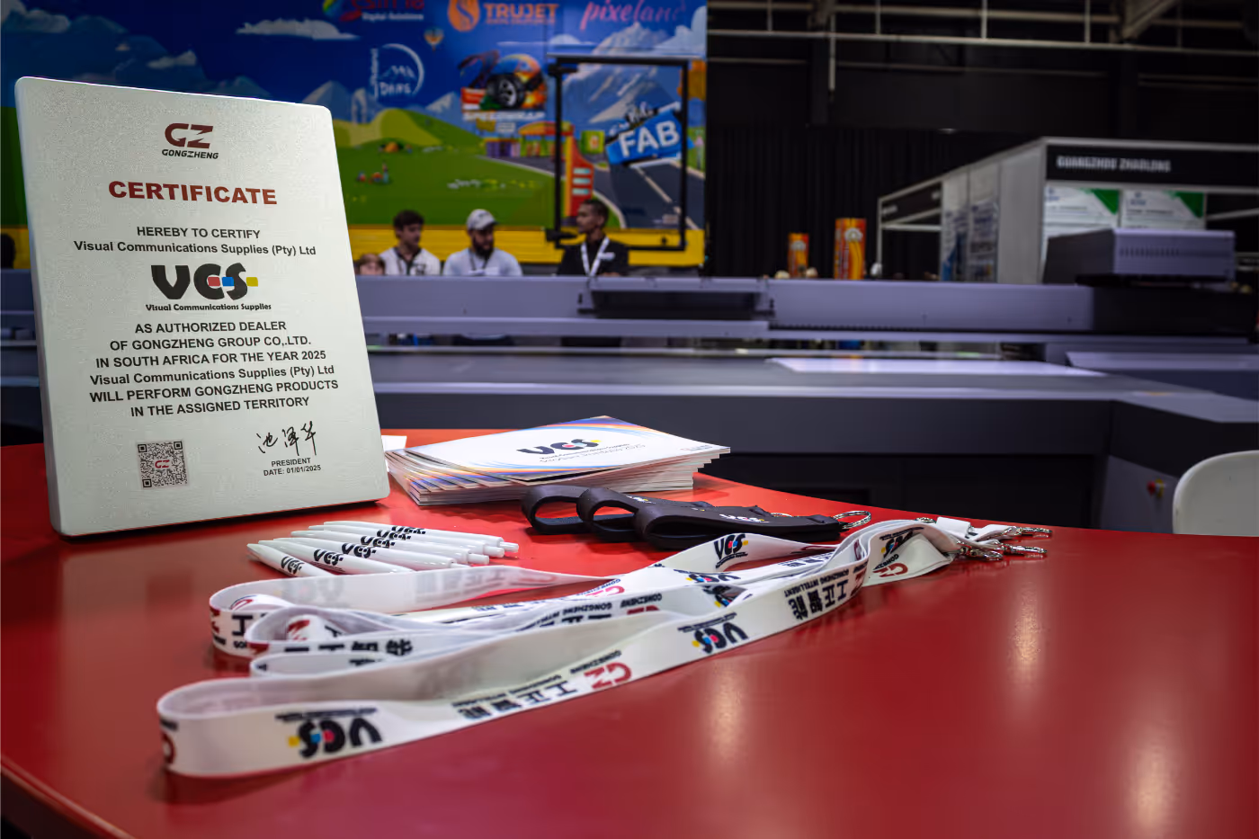 Certificate on a red table certifying Visual Communications Supplies as authorized dealer of Gongzheng Group in South Africa for 2025, with branded lanyards, pens, and brochures nearby.