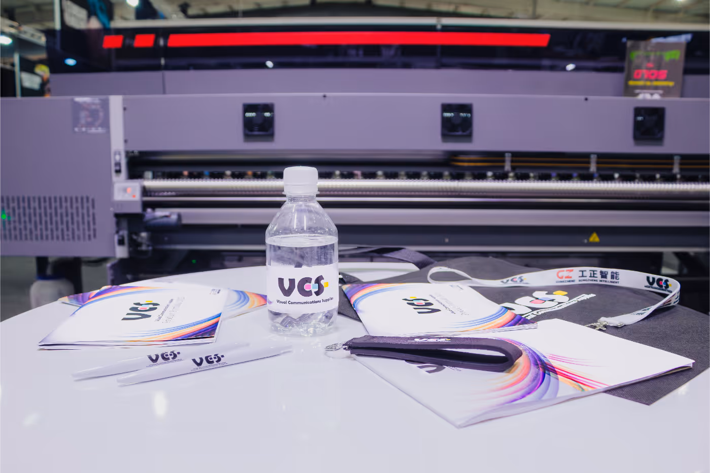 Table displaying branded promotional items including a water bottle, pens, notebooks, and a lanyard with VCS logo in front of a large industrial printer.