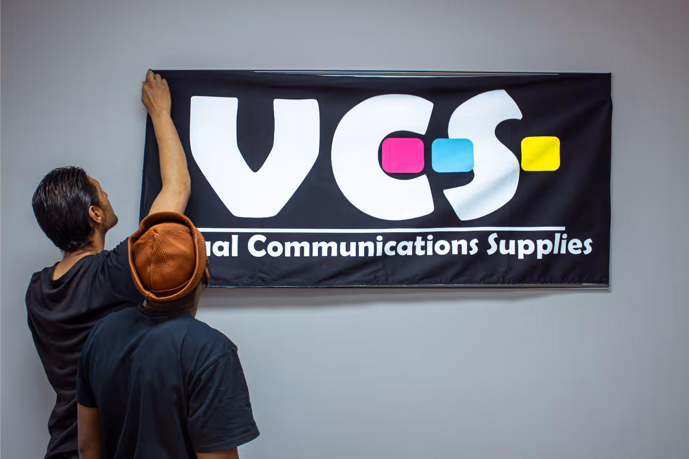 Two men hanging a black banner on a wall that reads 'VCS Visual Communications Supplies' with colorful blocks inside the letters.