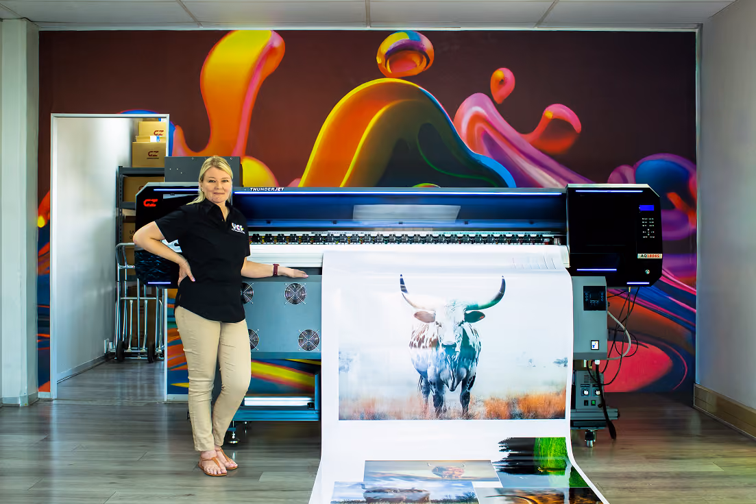 Woman standing next to a large format printer producing posters, including a prominent image of a bull with large horns, in a room with colorful abstract wall art.