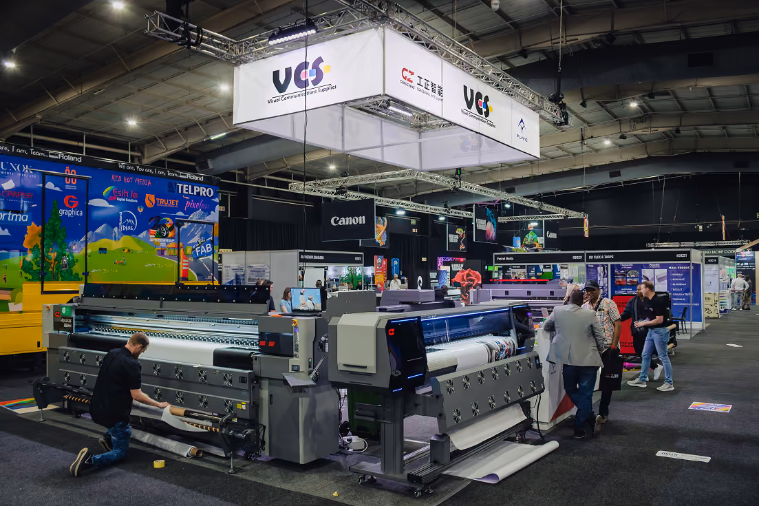 Indoor trade show or exhibition featuring large printing machines and equipment, with several people inspecting the machines under signage displaying company names including VCS and Canon.
