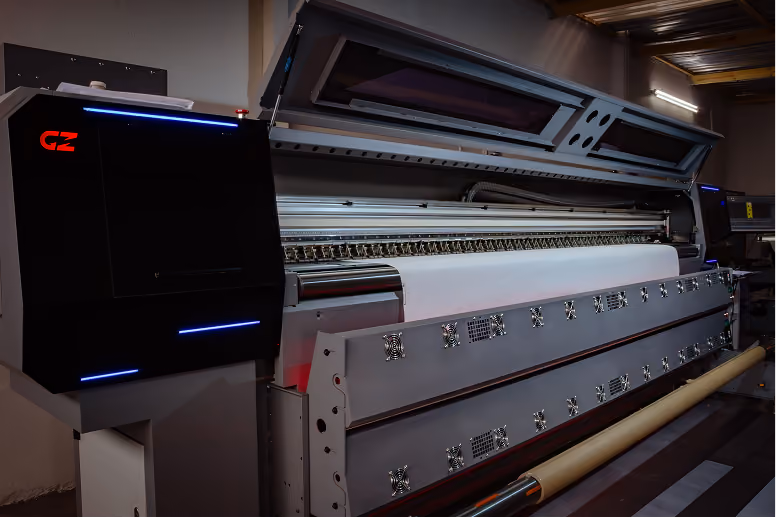Large industrial digital printer with an open top cover, printing on a wide roll of white material in a dimly lit room.