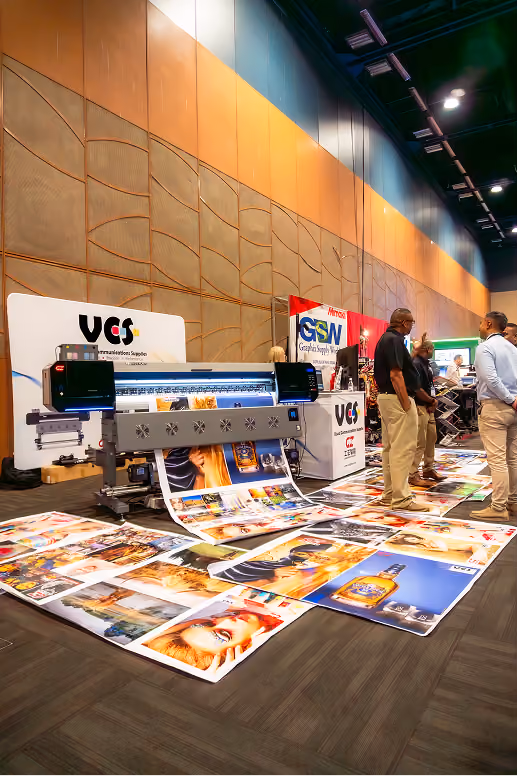 Large industrial printer producing colorful large-format prints spread out on the floor at a trade show, with several people standing nearby and company banners in the background.