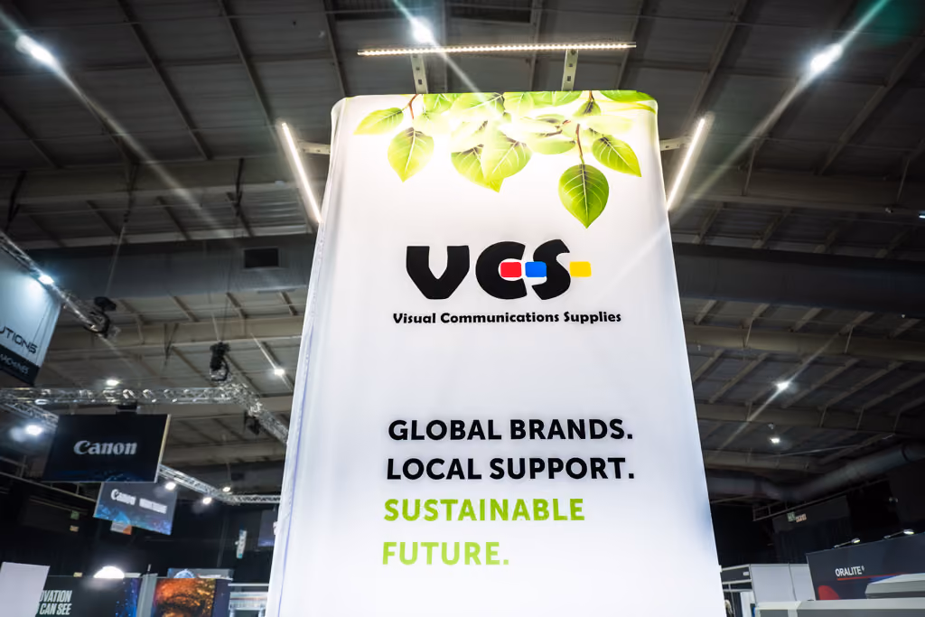 A tall white banner with green leaves at the top displaying the logo 'VCS Visual Communications Supplies' followed by the text 'GLOBAL BRANDS. LOCAL SUPPORT. SUSTAINABLE FUTURE.' in black and green letters inside a convention or exhibition hall with visible lighting and Canon signage in the background.