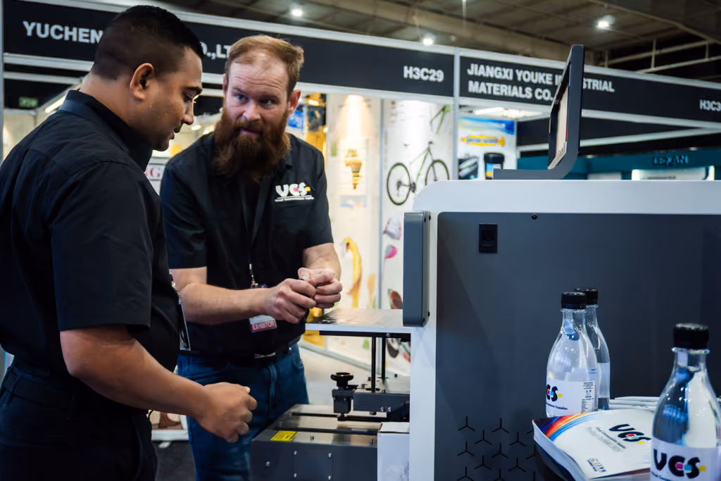 Two men in black shirts engaged in discussion next to a printing machine at an industrial trade booth displaying brochures and water bottles labeled UCS.