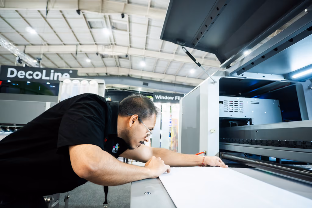 Man leaning over large industrial printing machine, adjusting material on the printer bed inside a well-lit manufacturing or trade show environment.