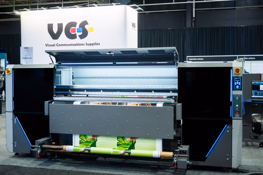Large industrial printer producing a wide-format print with a green-themed image, displayed in an exhibition setting with a white sign behind reading 'VCS Visual Communications Supplies'.