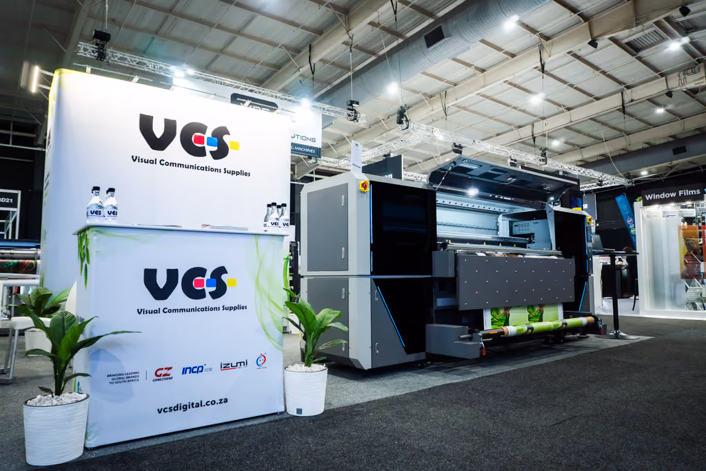 Exhibition booth for VCS Visual Communications Supplies featuring a large digital printer printing colorful material, a display counter with VCS branding, four water bottles with VCS logo, two potted plants, and bright overhead lights in an indoor event space.
