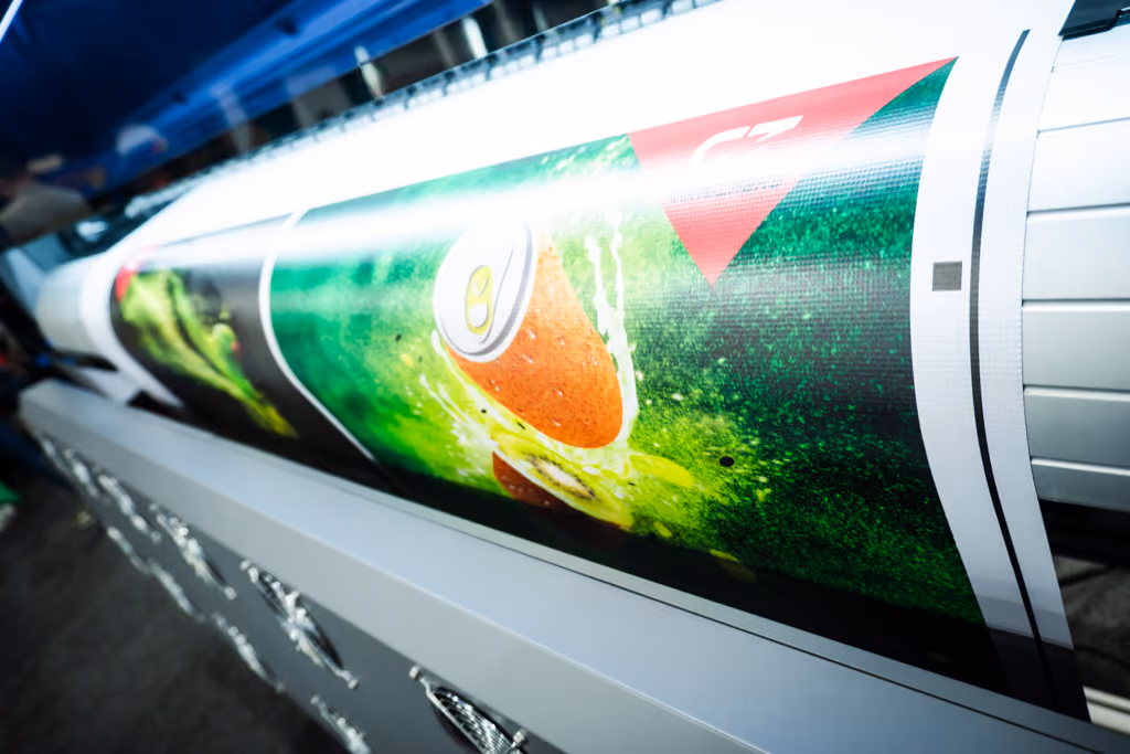 Large printer producing a vibrant print featuring an orange soda can splashing into green liquid with red and white triangular design elements.