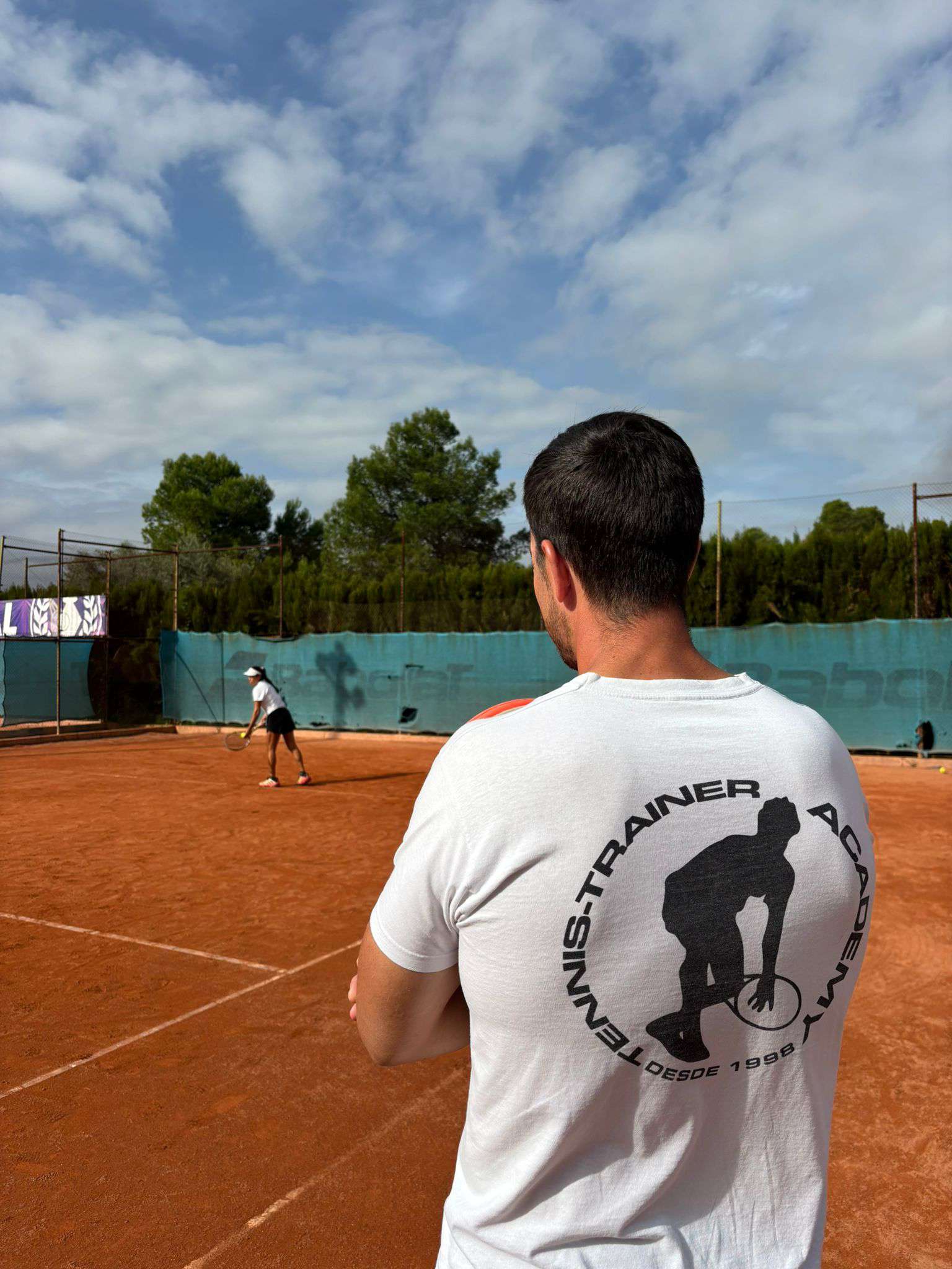 Tennis coach in a white shirt with a tennis player logo on the back watching a player prepare to serve on a clay tennis court.