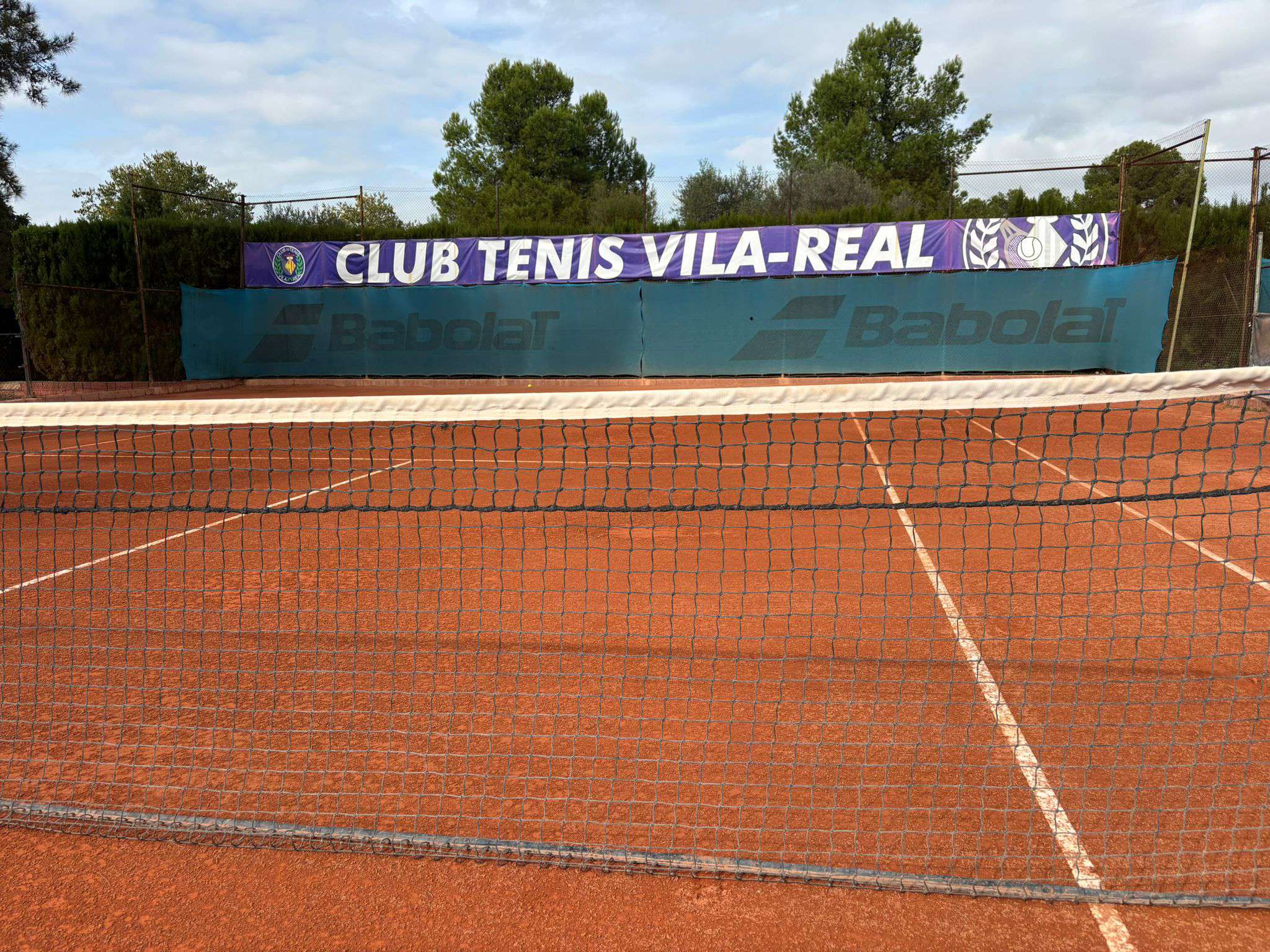 Empty clay tennis court with a net in the foreground and a Club Tenis Vila-Real banner in the background.
