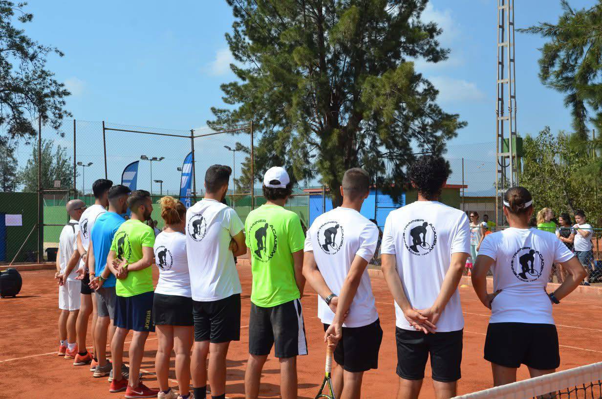 Group of tennis players standing in a line on a clay court with their backs to the camera, wearing white and neon green shirts with a tennis trainer logo.