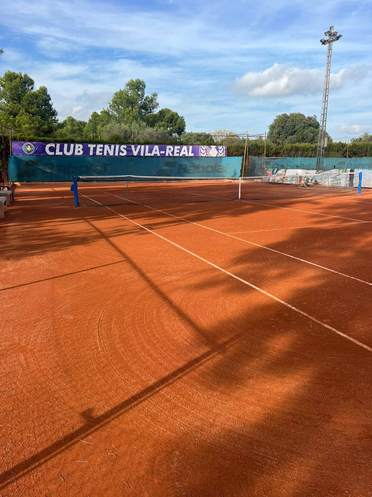 Empty clay tennis court with net, surrounded by green trees and blue sky, at Club Tenis Vila-Real.