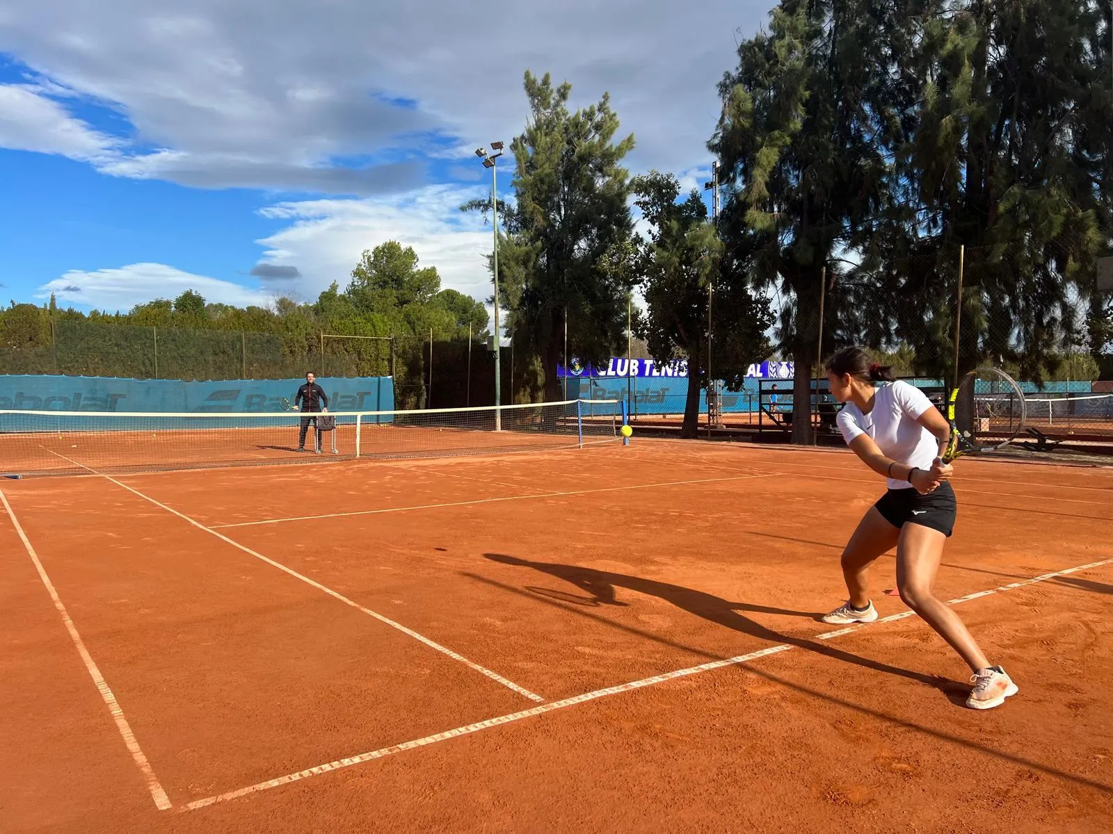 Woman in white shirt and black shorts preparing to hit a tennis ball on an outdoor clay court with a man standing on the opposite side.