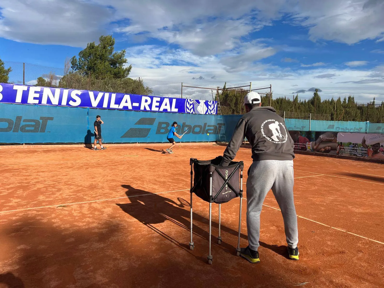 Tennis coach standing with ball hopper on clay court while two players practice returning a ball.