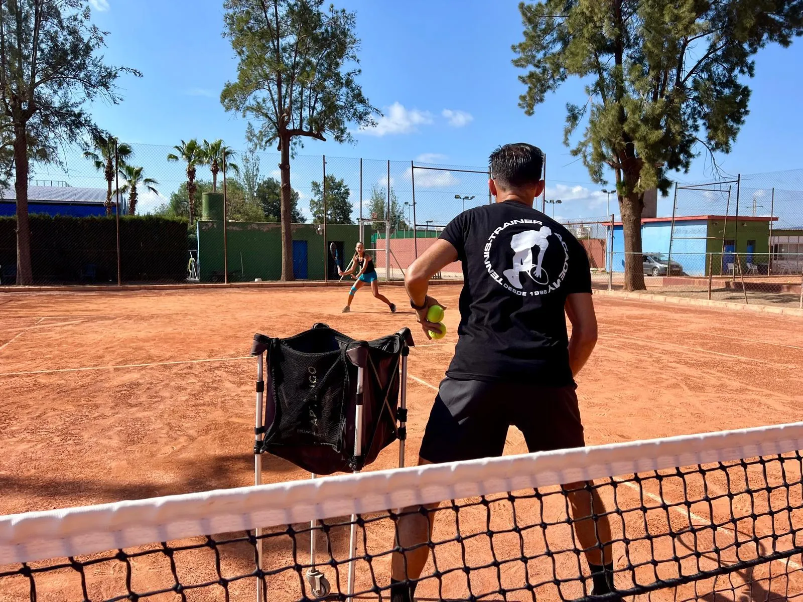 Tennis coach with balls in hand facing a player ready to hit on a clay court during a training session.