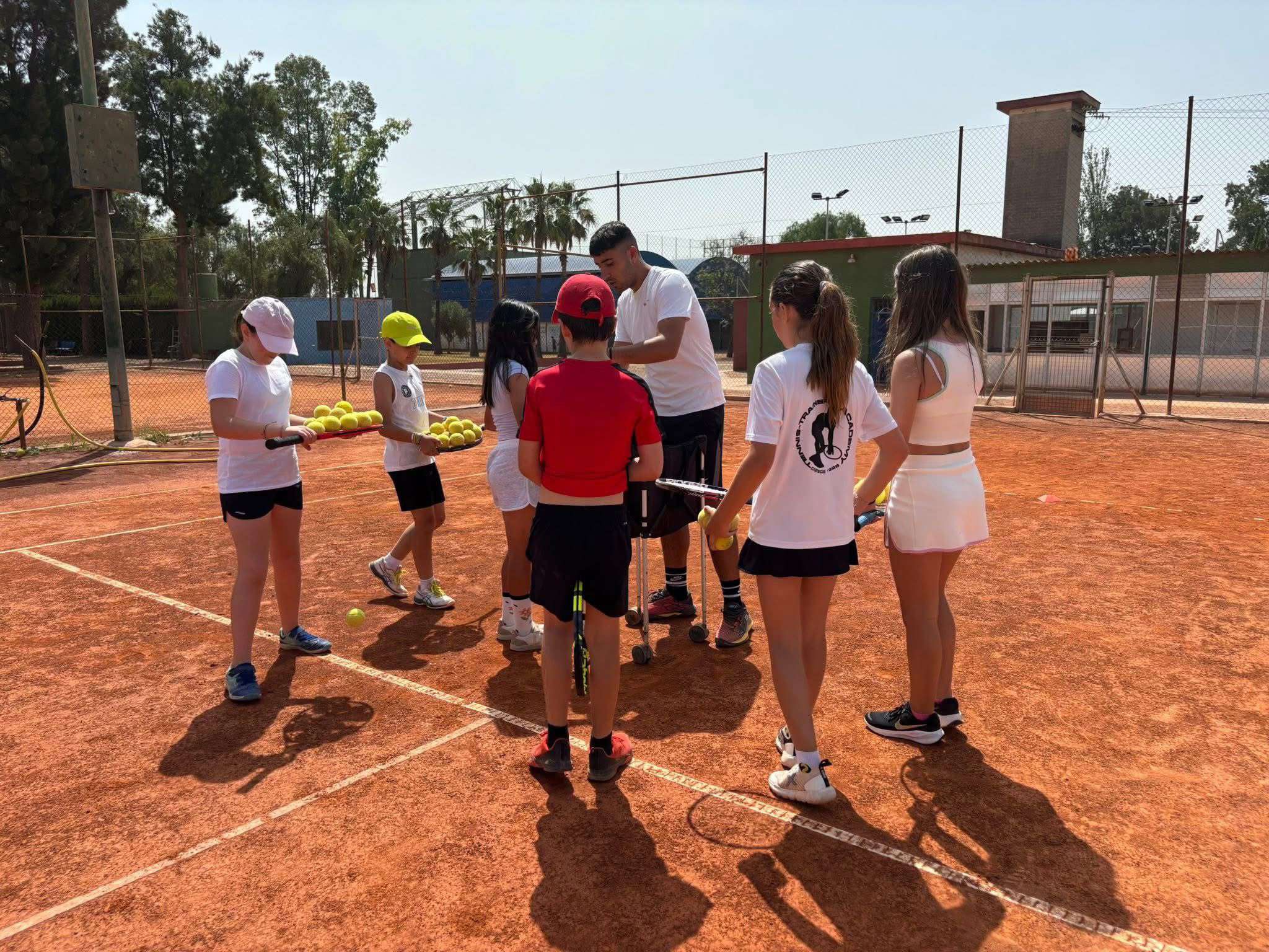 A tennis coach instructs five young players holding tennis balls and rackets on a clay tennis court.