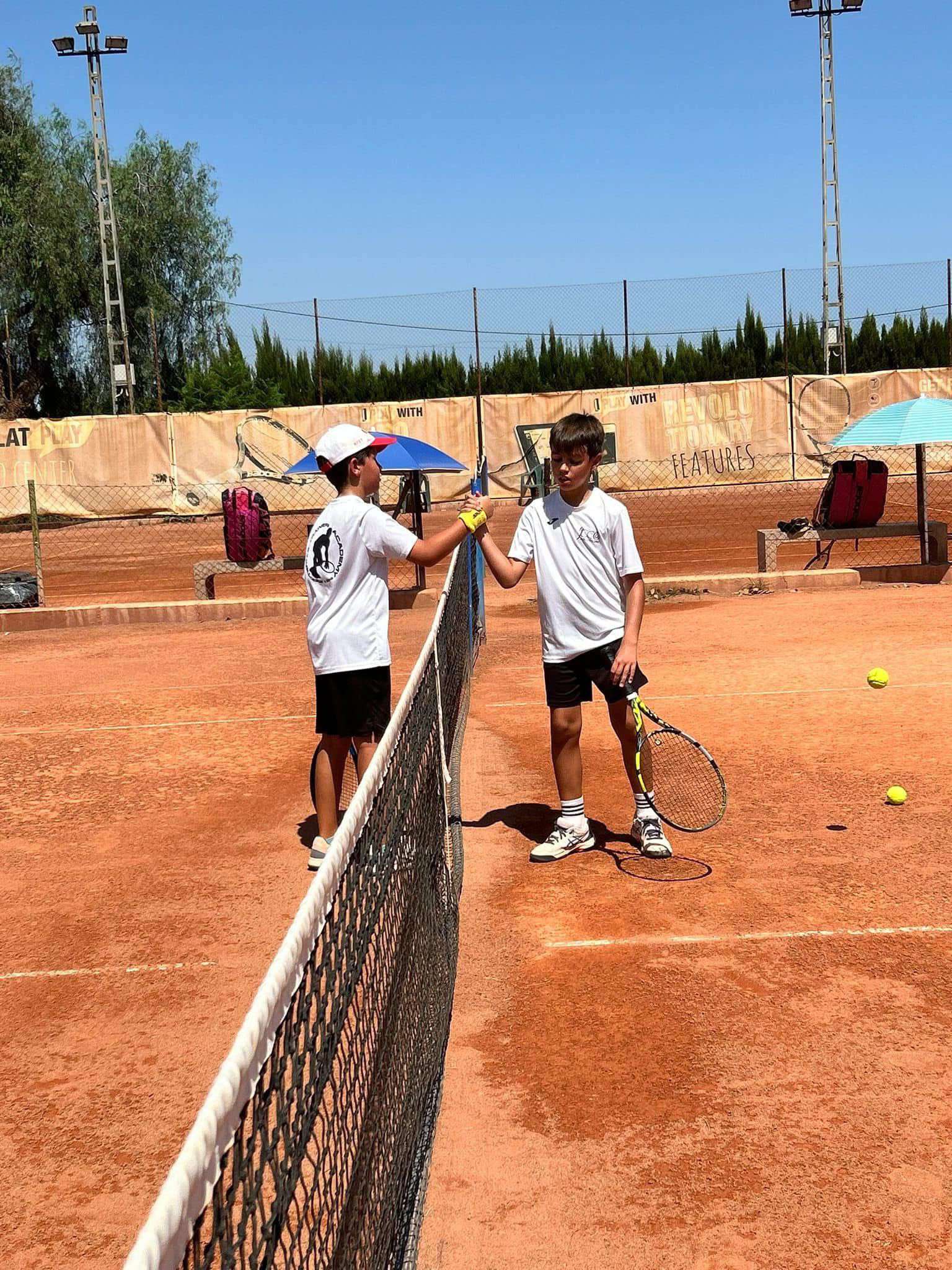 Two boys in white shirts and black shorts fist bumping over a tennis net on a clay court with tennis balls scattered nearby.