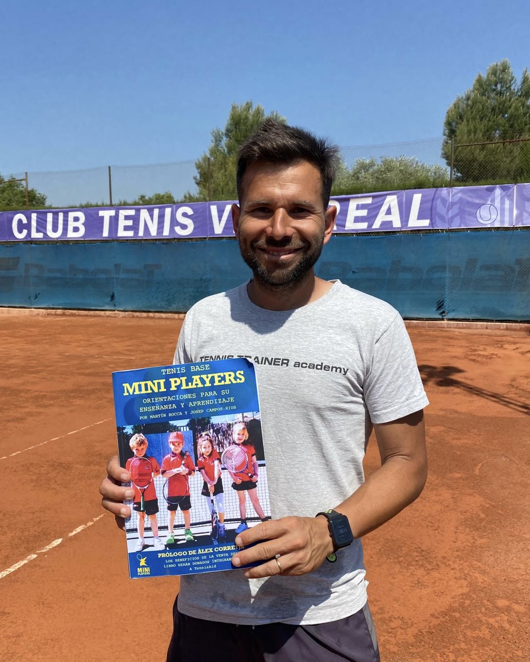 Man smiling on a clay tennis court holding a book titled 'Mini Players' with images of children holding tennis rackets.