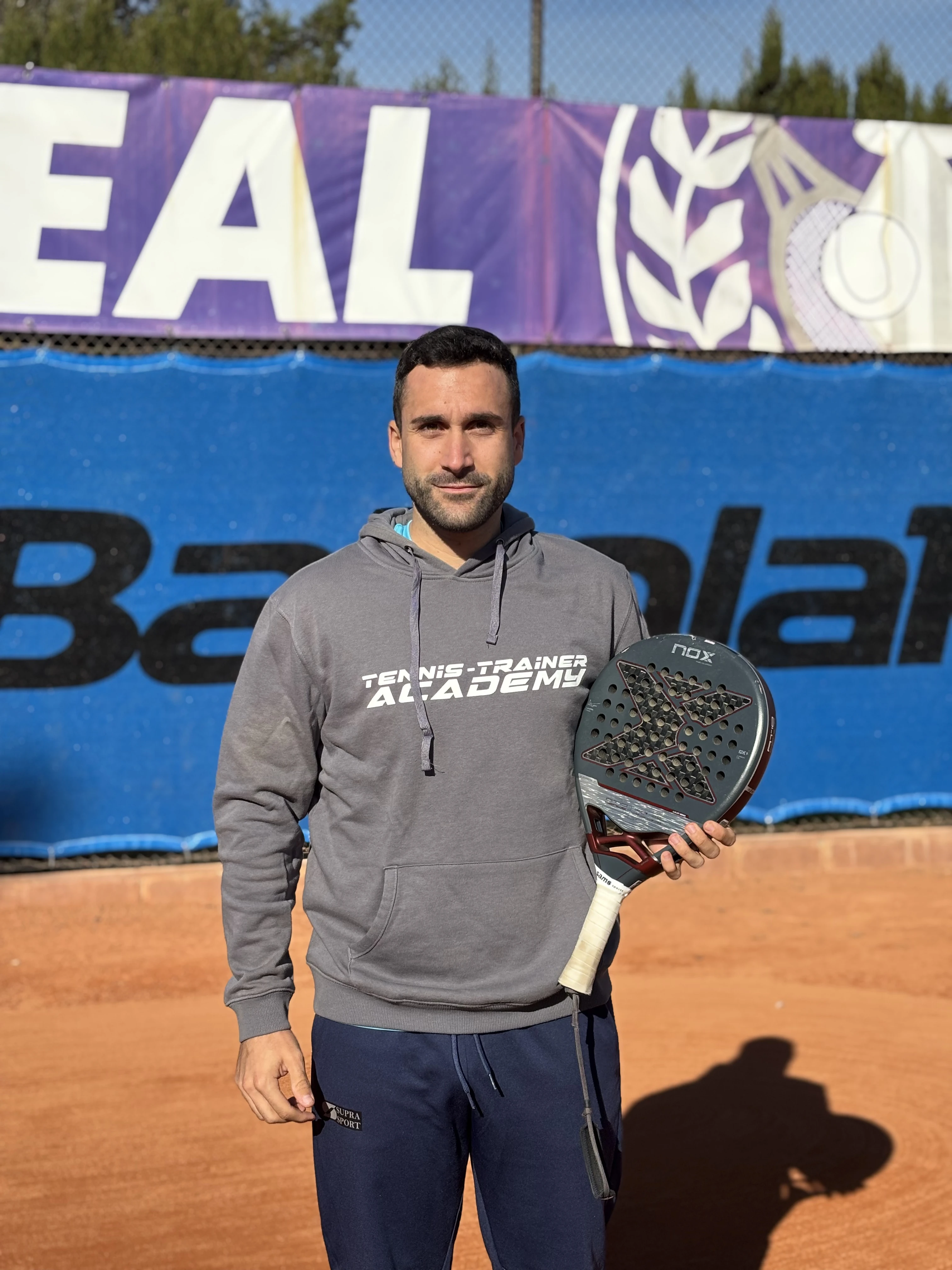 Man in gray hoodie holding a padel racket on a clay court with a banner in the background.