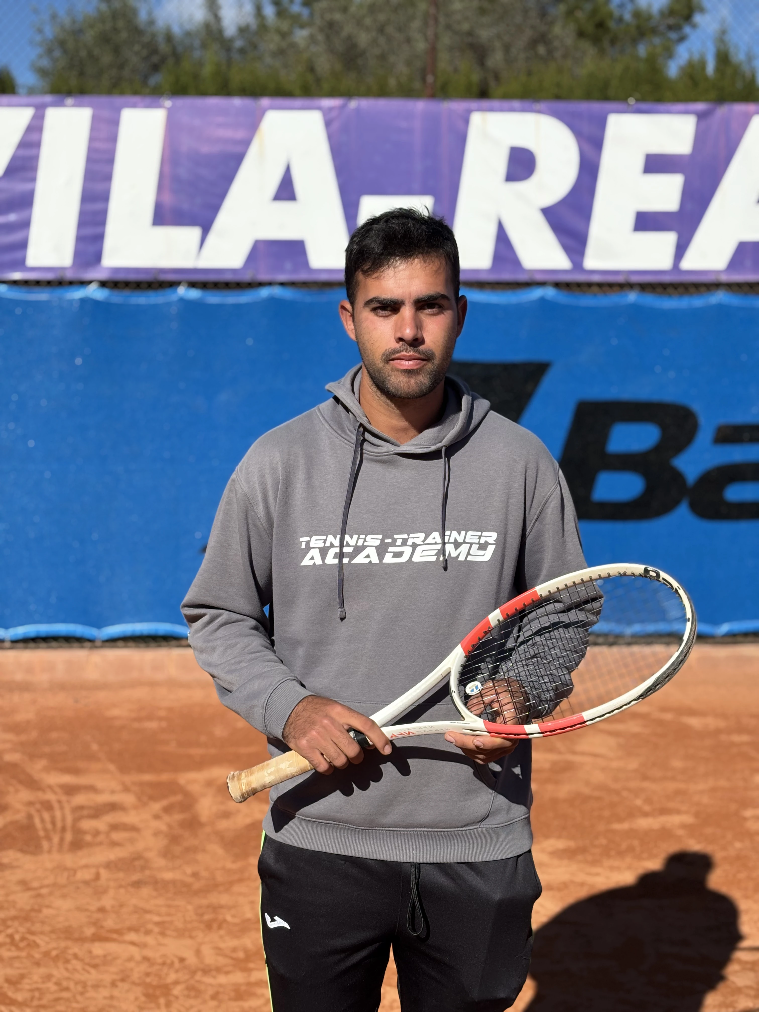 Man in gray hoodie holding a tennis racket on a clay tennis court with a large blue and purple banner in the background.