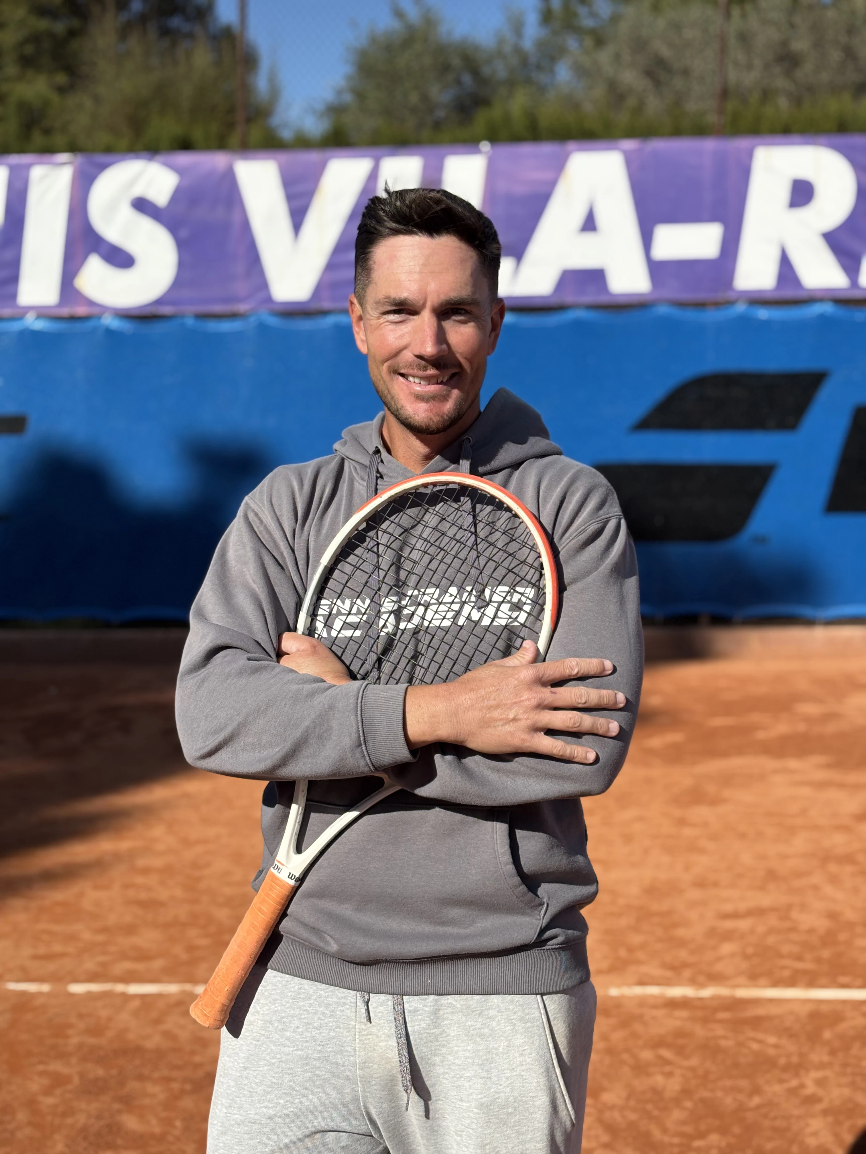 Smiling man in gray hoodie holding a tennis racquet crossed over his chest on a clay tennis court.
