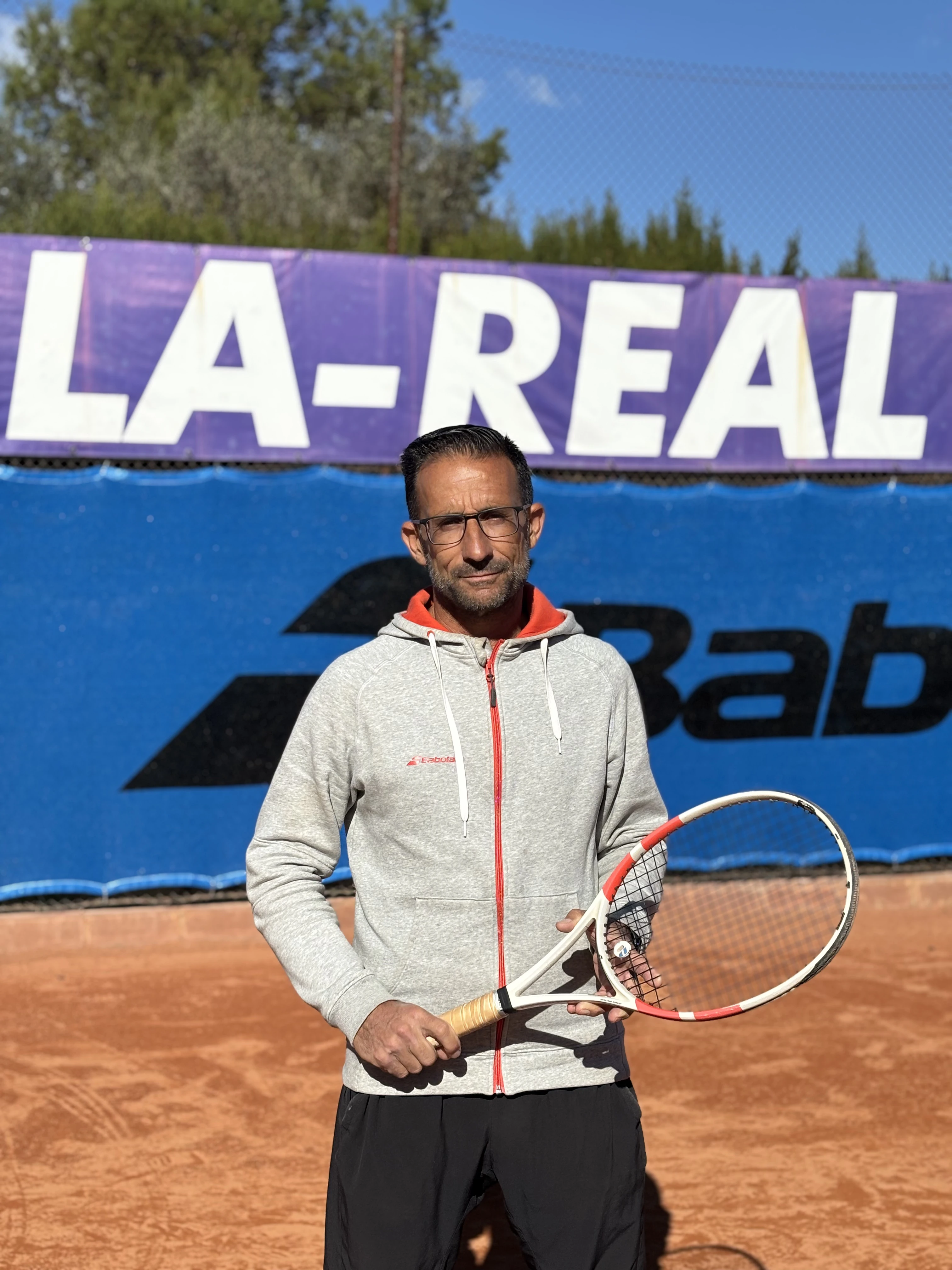 Man wearing glasses and a gray hoodie holding a tennis racket on a clay tennis court with banners in the background.