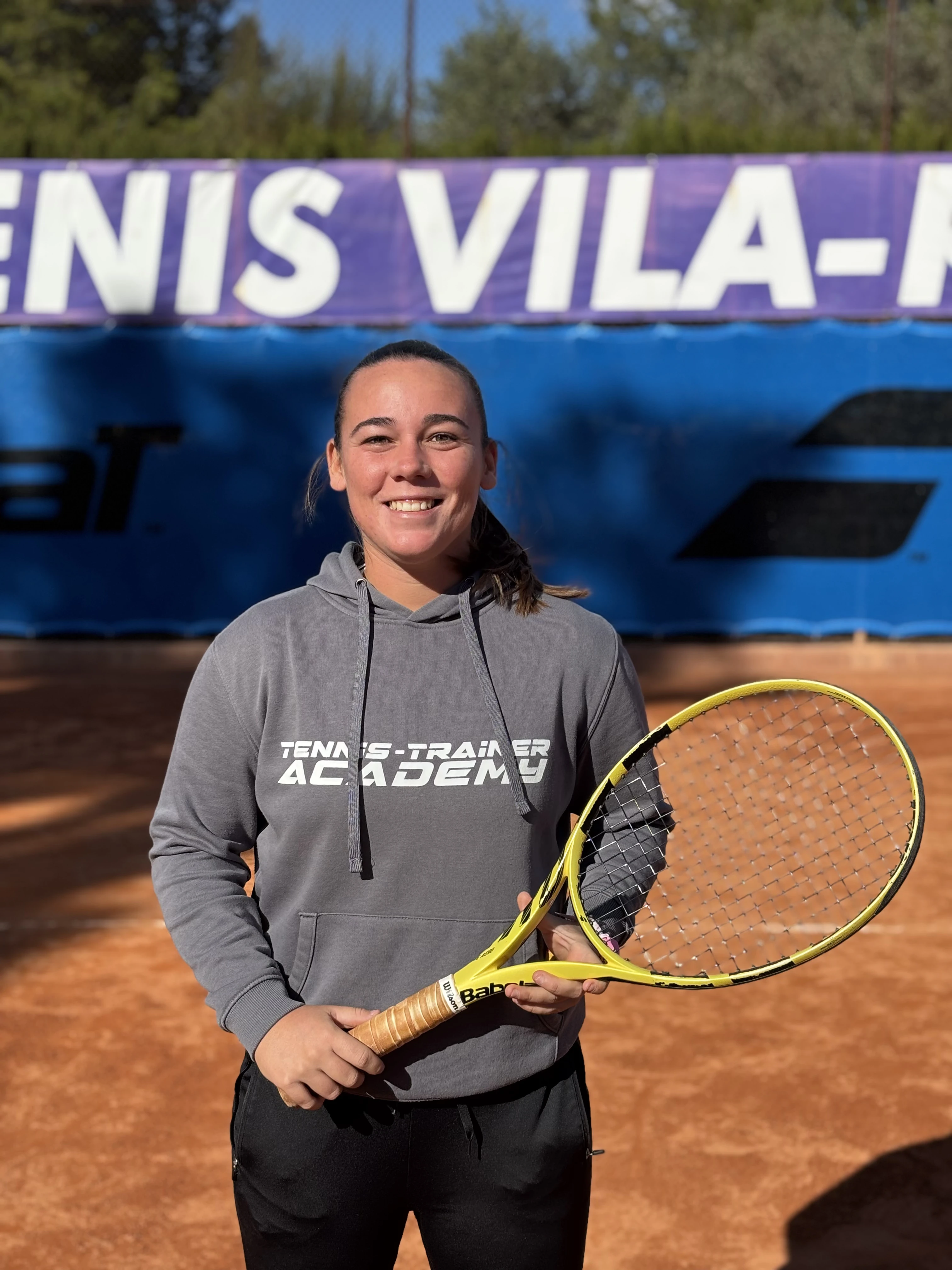 Smiling woman holding a yellow tennis racket on a clay tennis court with a banner in the background.