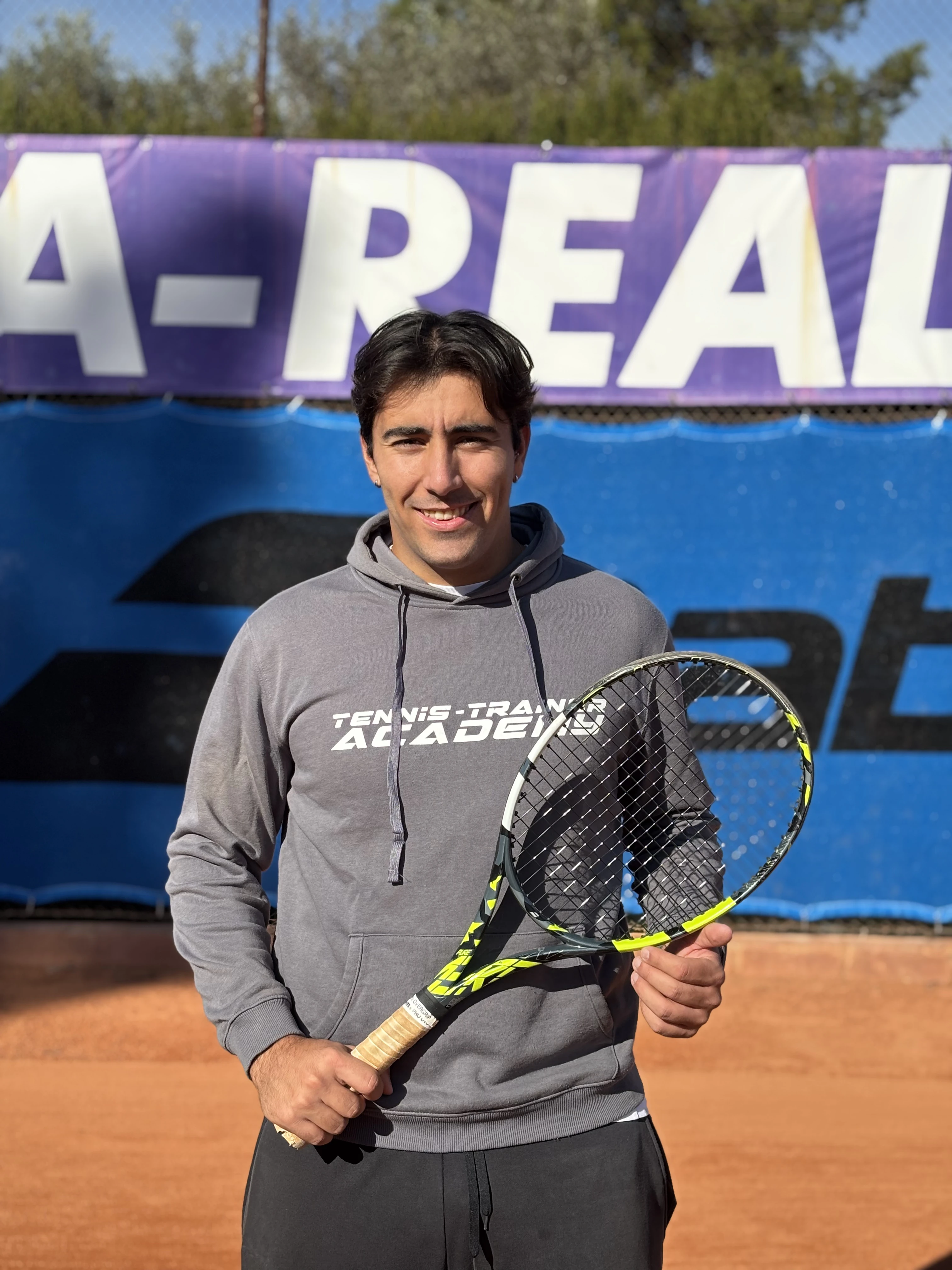 Smiling young man in a gray hoodie holding a tennis racket on a clay tennis court.