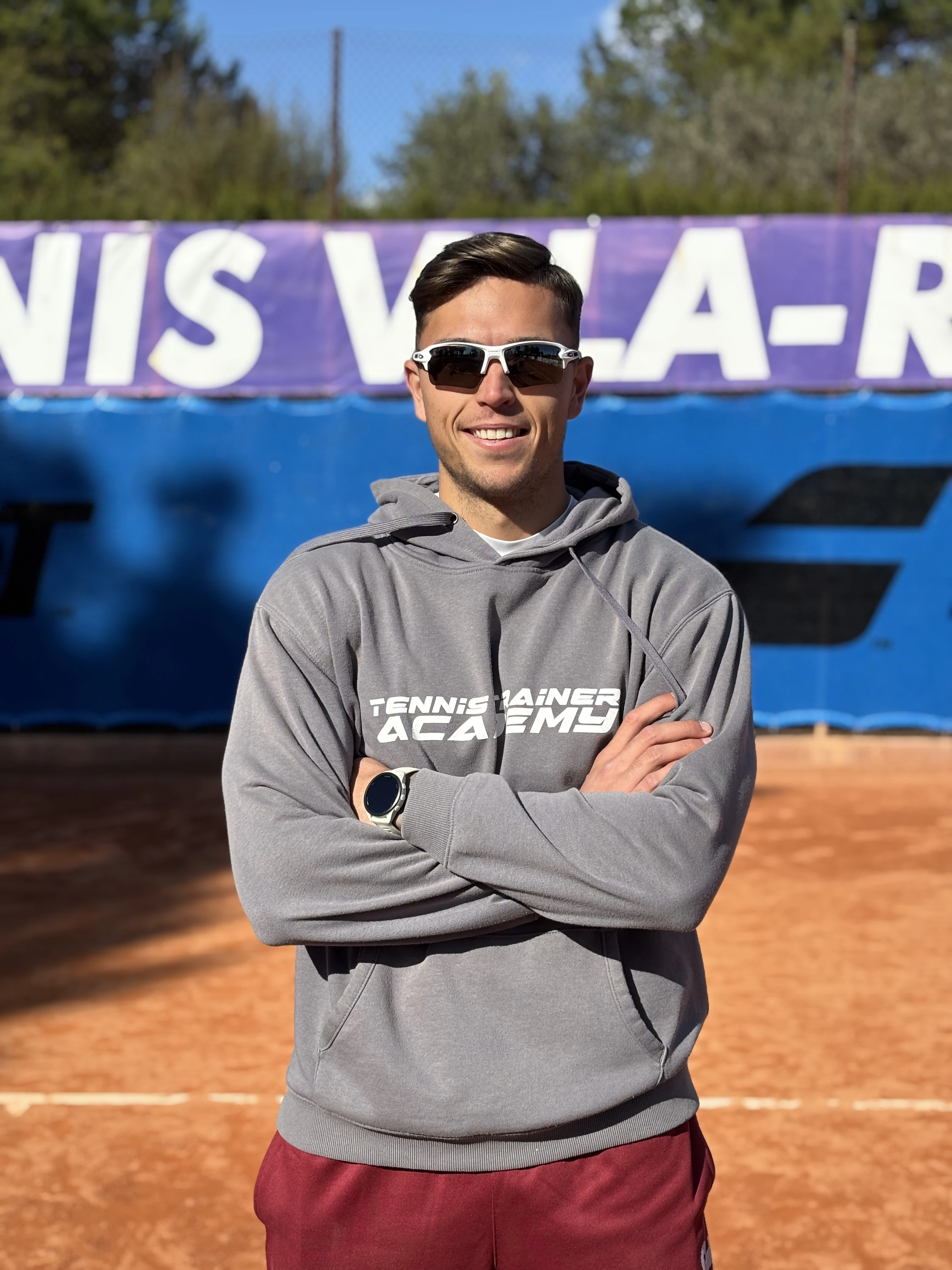 Smiling man wearing white sports sunglasses and grey hoodie with 'Tennis Trainer Academy' text standing on clay tennis court with arms crossed.