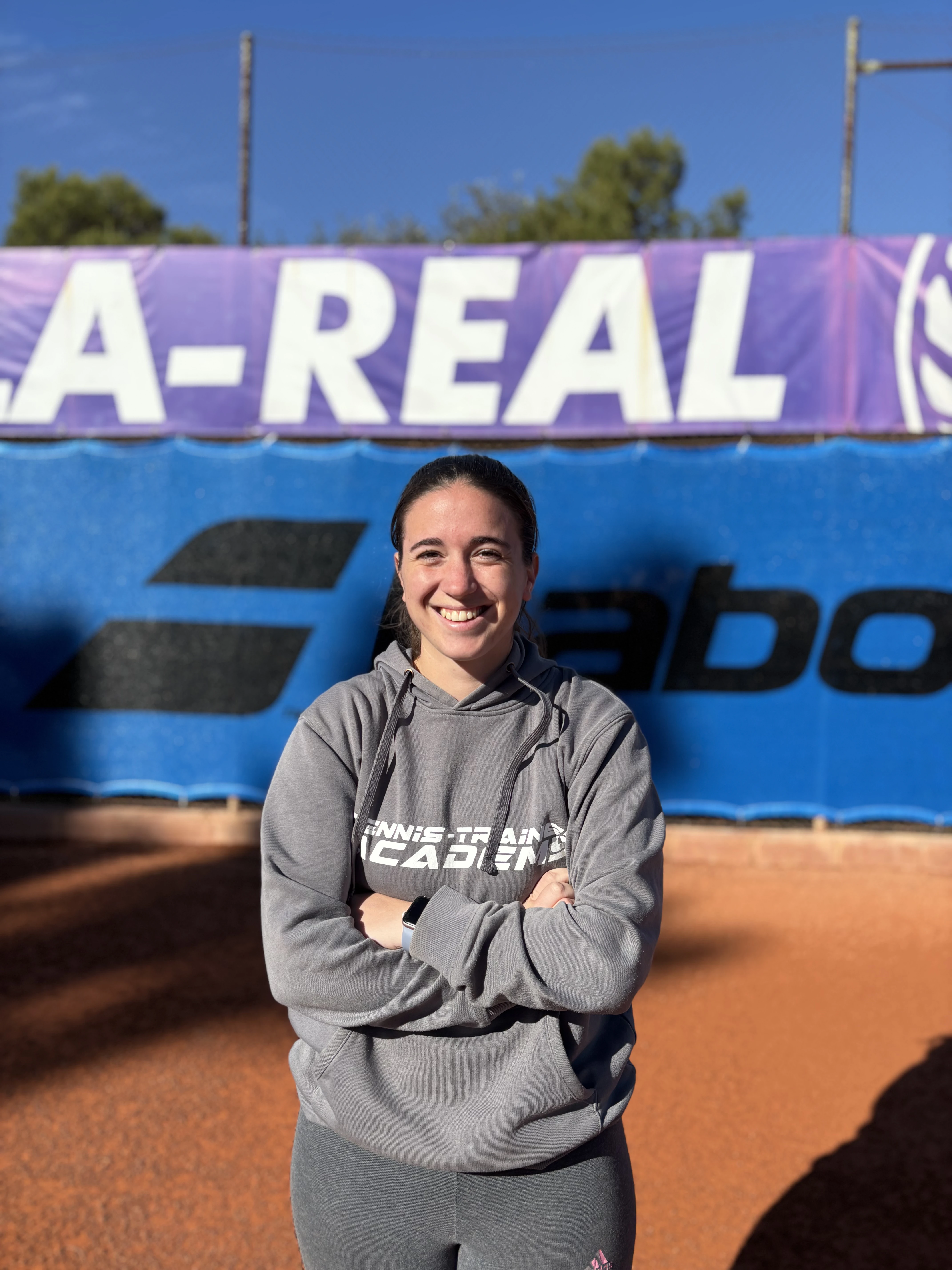 Smiling woman in gray hoodie and leggings standing on a clay tennis court with advertising banners in the background.