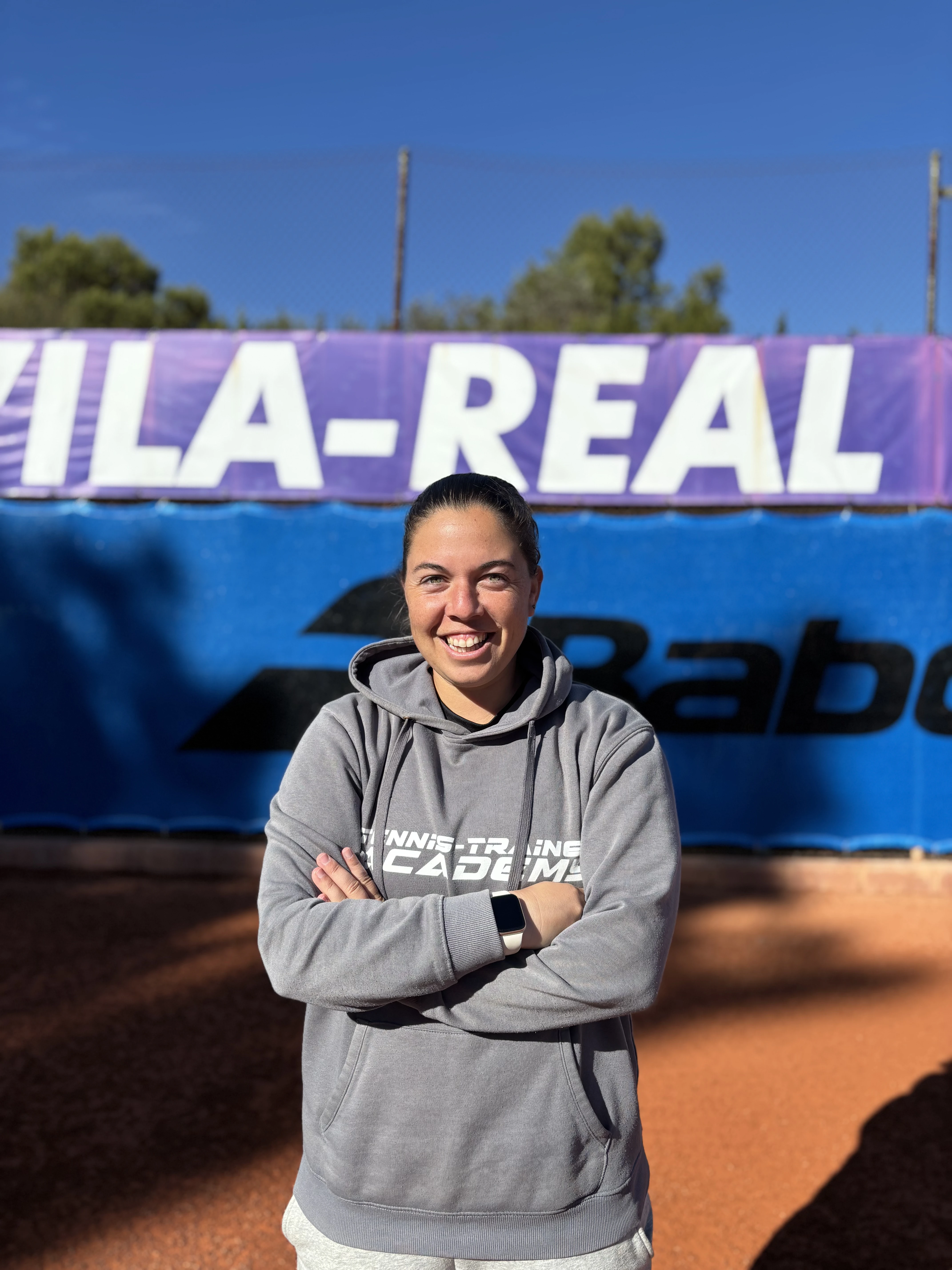 Smiling woman in a gray hoodie with crossed arms standing on a tennis court with a purple and blue banner in the background.
