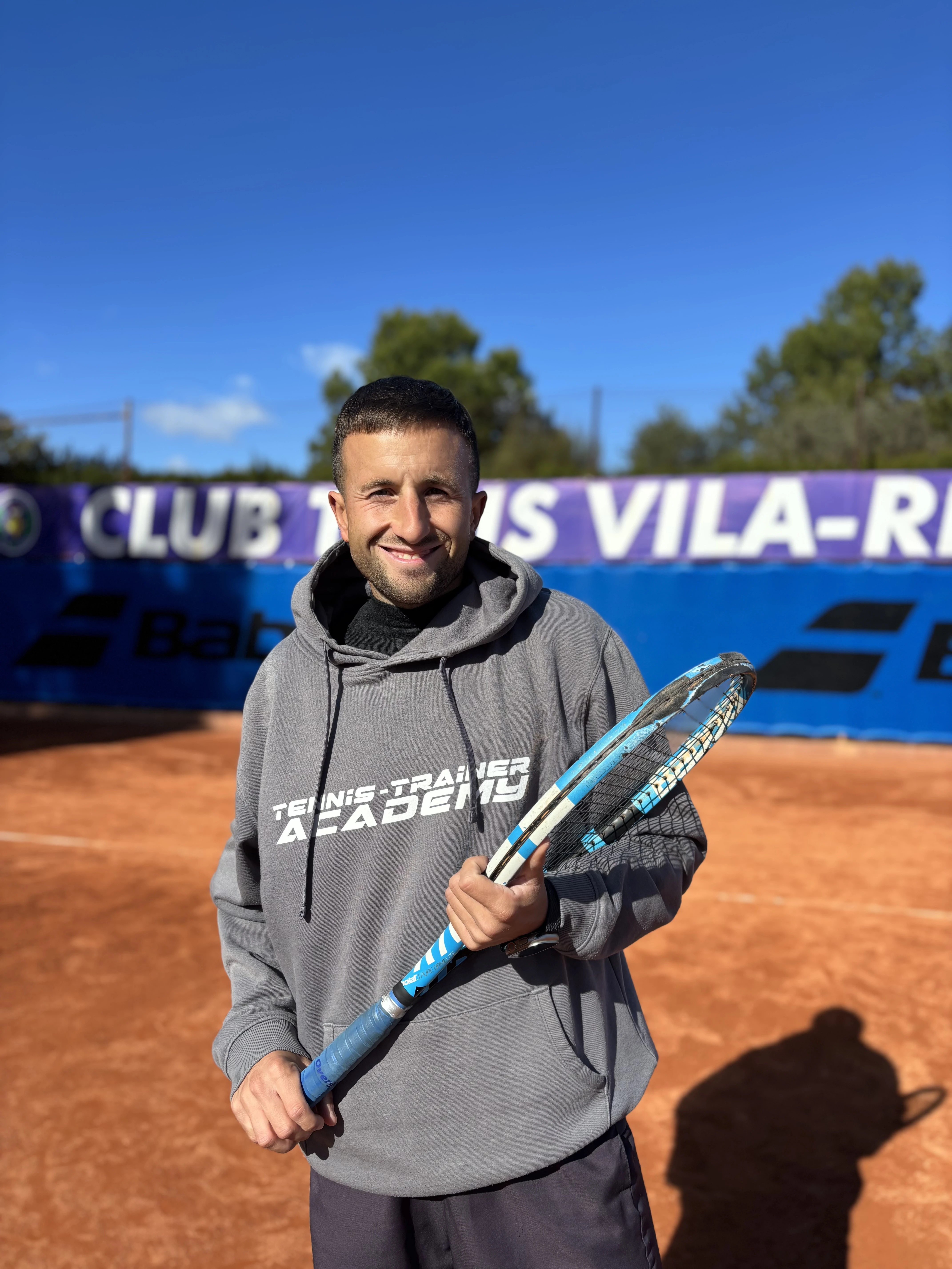 Smiling man in gray hoodie holding a blue tennis racket on an outdoor clay tennis court under a clear blue sky.