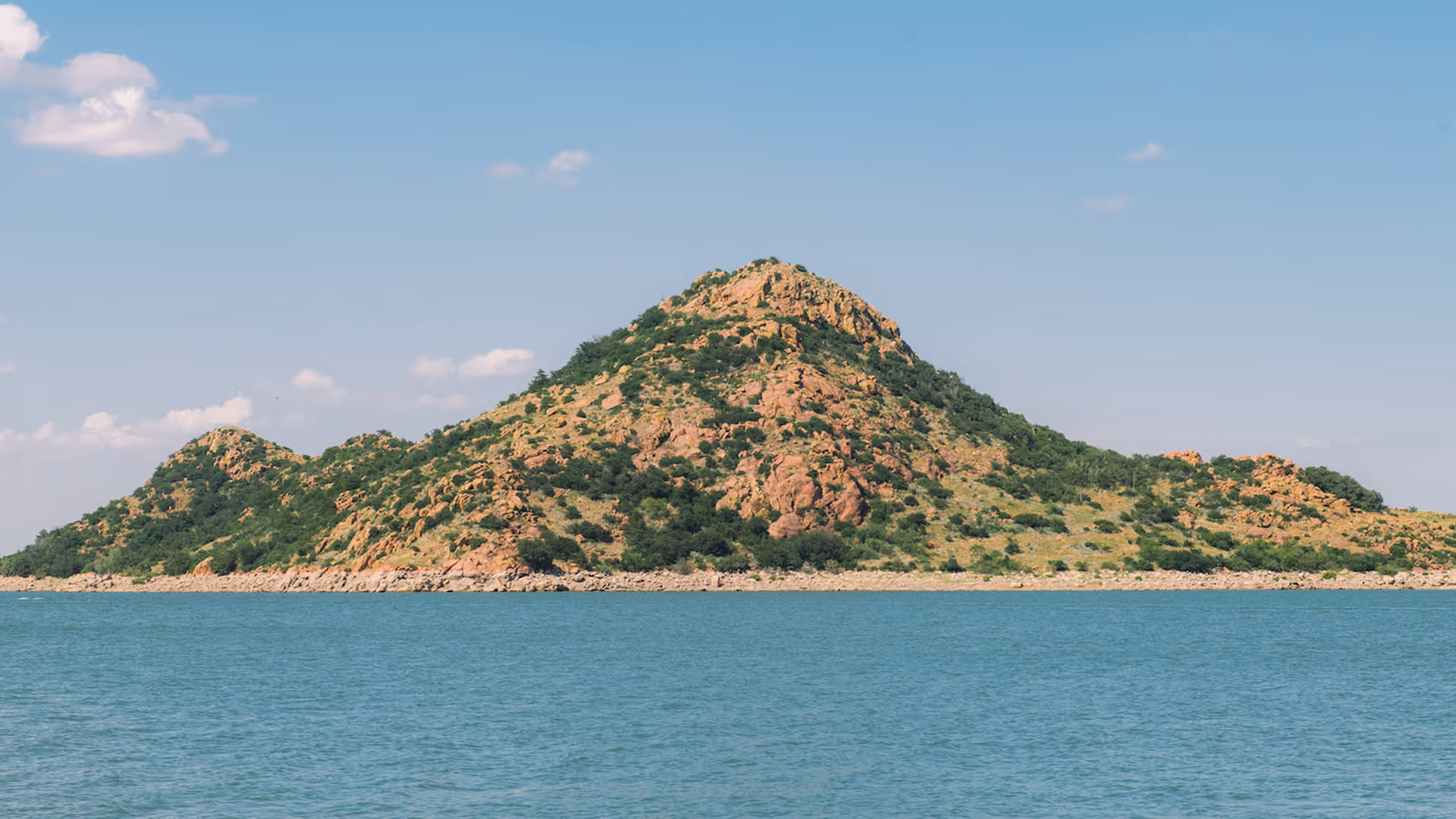 Rocky island with scattered greenery surrounded by blue water under a clear sky.