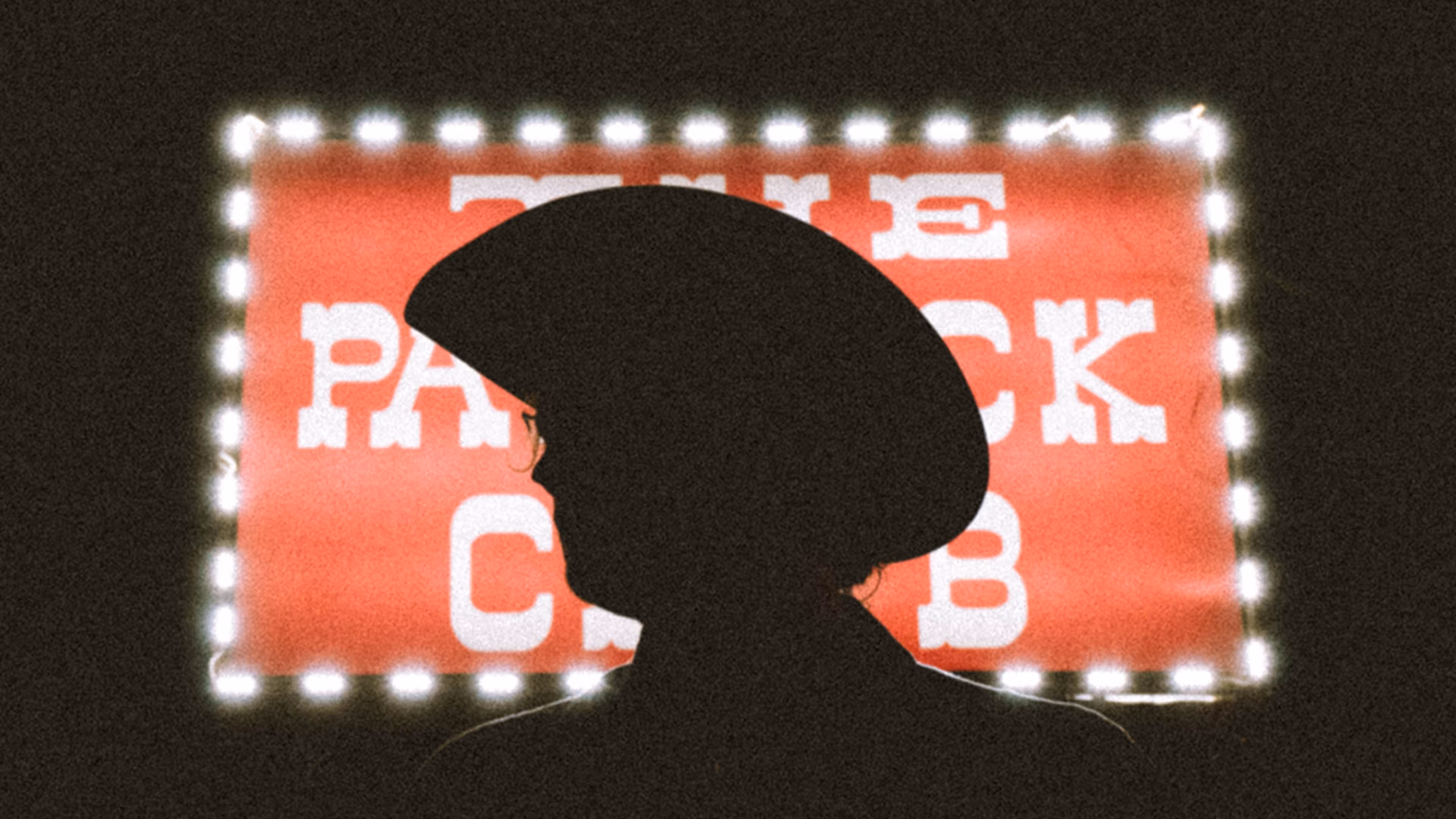 Silhouette of a person wearing a large hat and glasses in front of a lit sign reading 'THE PACK CLUB'.
