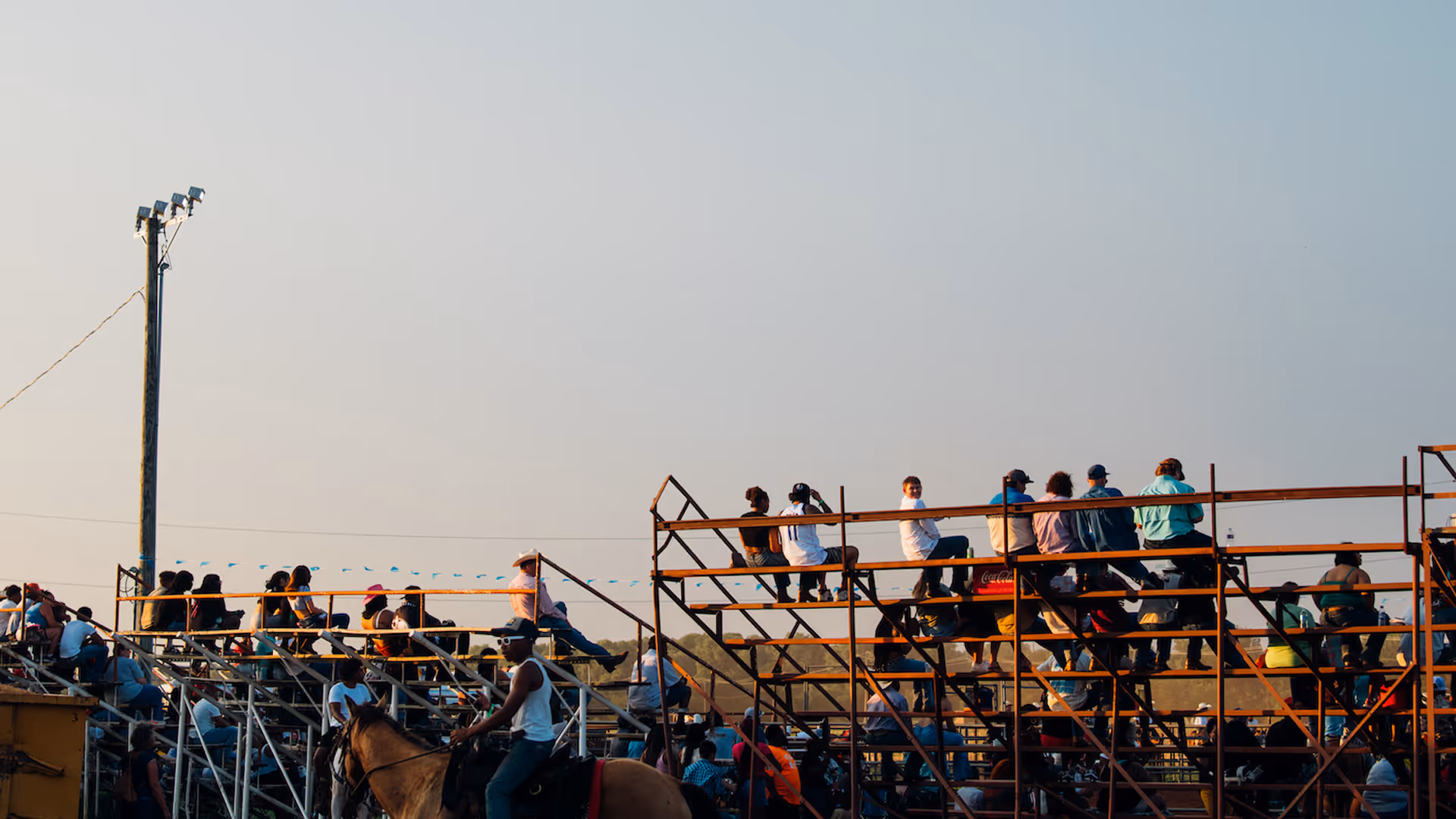 People sitting on metal bleachers outdoors during daytime, with one person riding a horse in the foreground.
