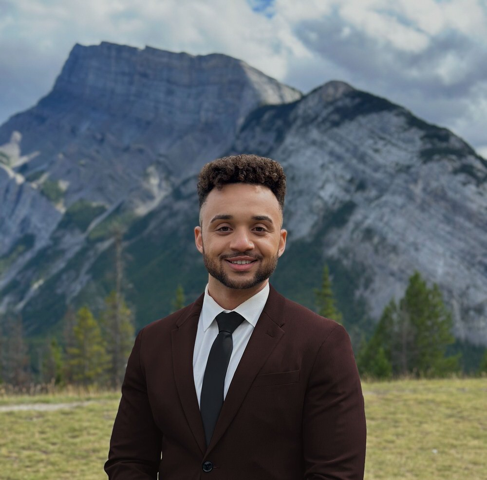 Smiling man with curly hair wearing a dark suit, white shirt, and black tie standing outdoors in front of a mountain landscape.